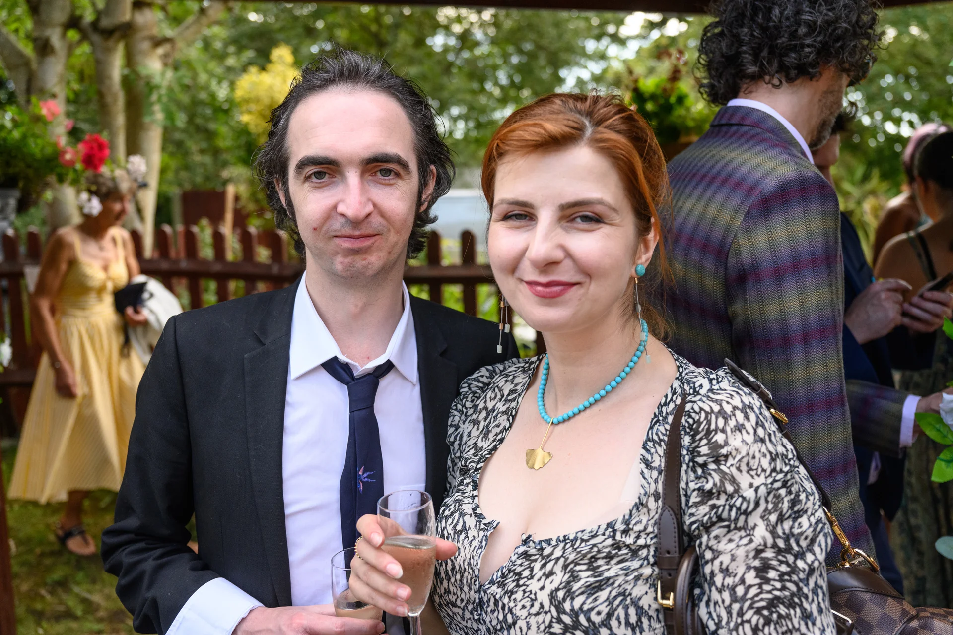 A smiling couple mingling outdoors at a wedding reception, holding glasses of champagne while guests chat in the background.