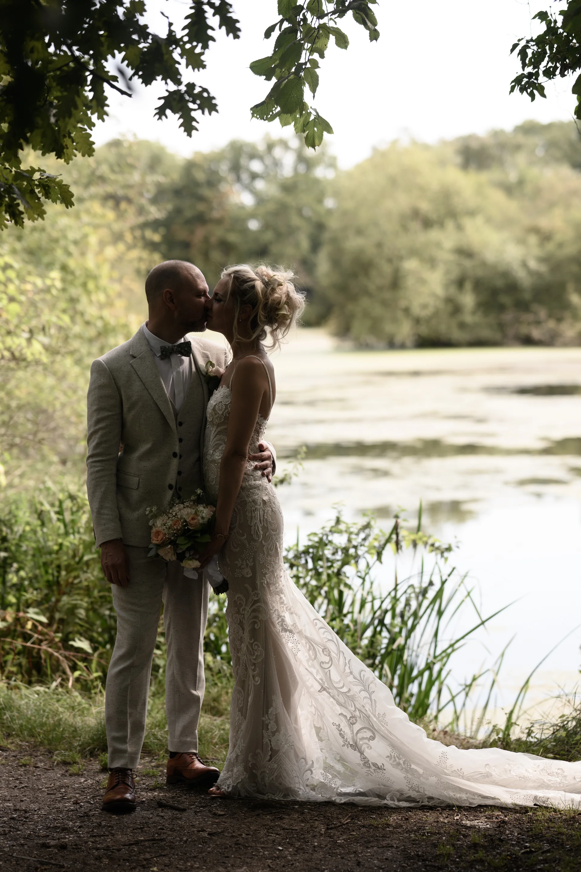 Newlyweds share a romantic kiss by a serene lake framed by overhanging tree branches during their outdoor portrait session.