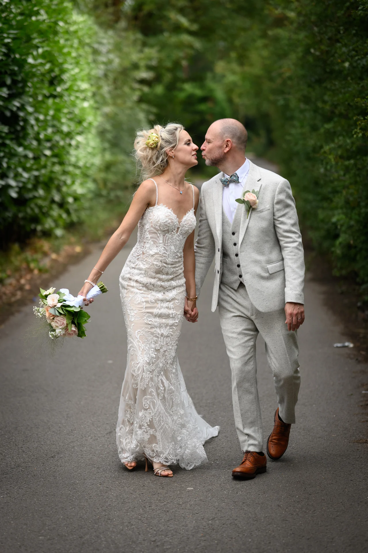 A bride in a lace fit-and-flare gown and groom in a light stone suit walk hand in hand down a tree-lined path, leaning in close for a tender moment.