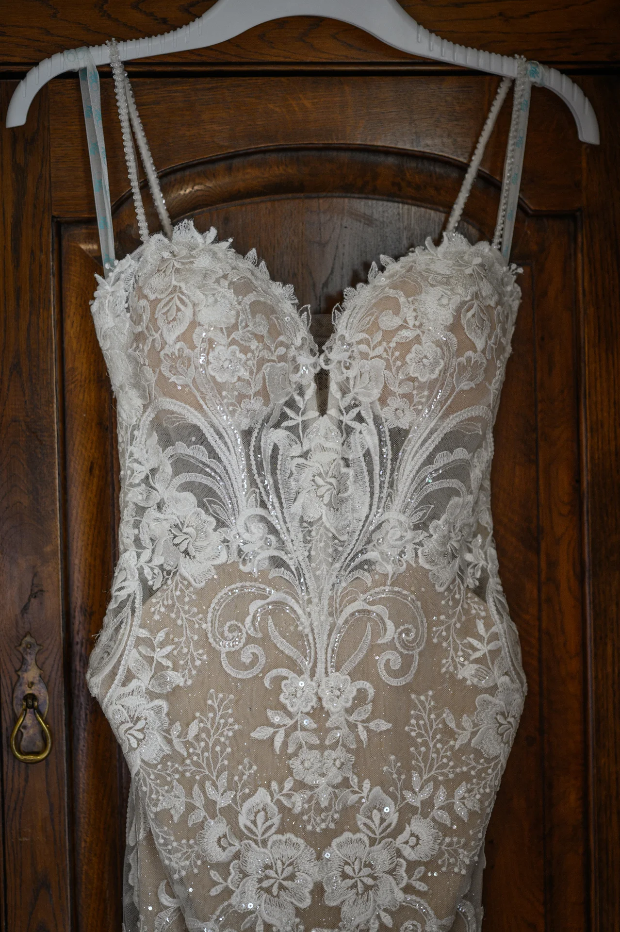 A stunning lace wedding dress featuring a plunging sweetheart neckline and delicate spaghetti straps hangs neatly against a rustic wooden wardrobe.
