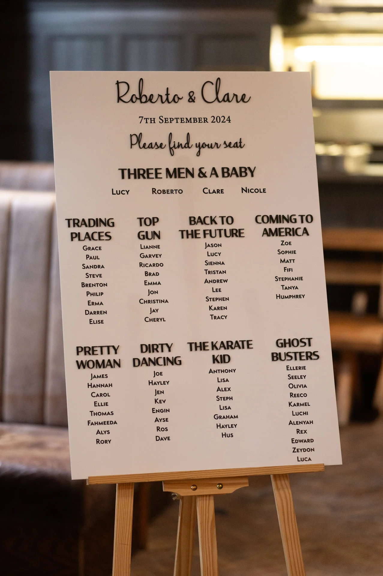 A modern white acrylic seating plan displayed on a wooden easel, featuring movie-themed table names like Top Gun and Ghostbusters for Roberto and Clare's wedding.