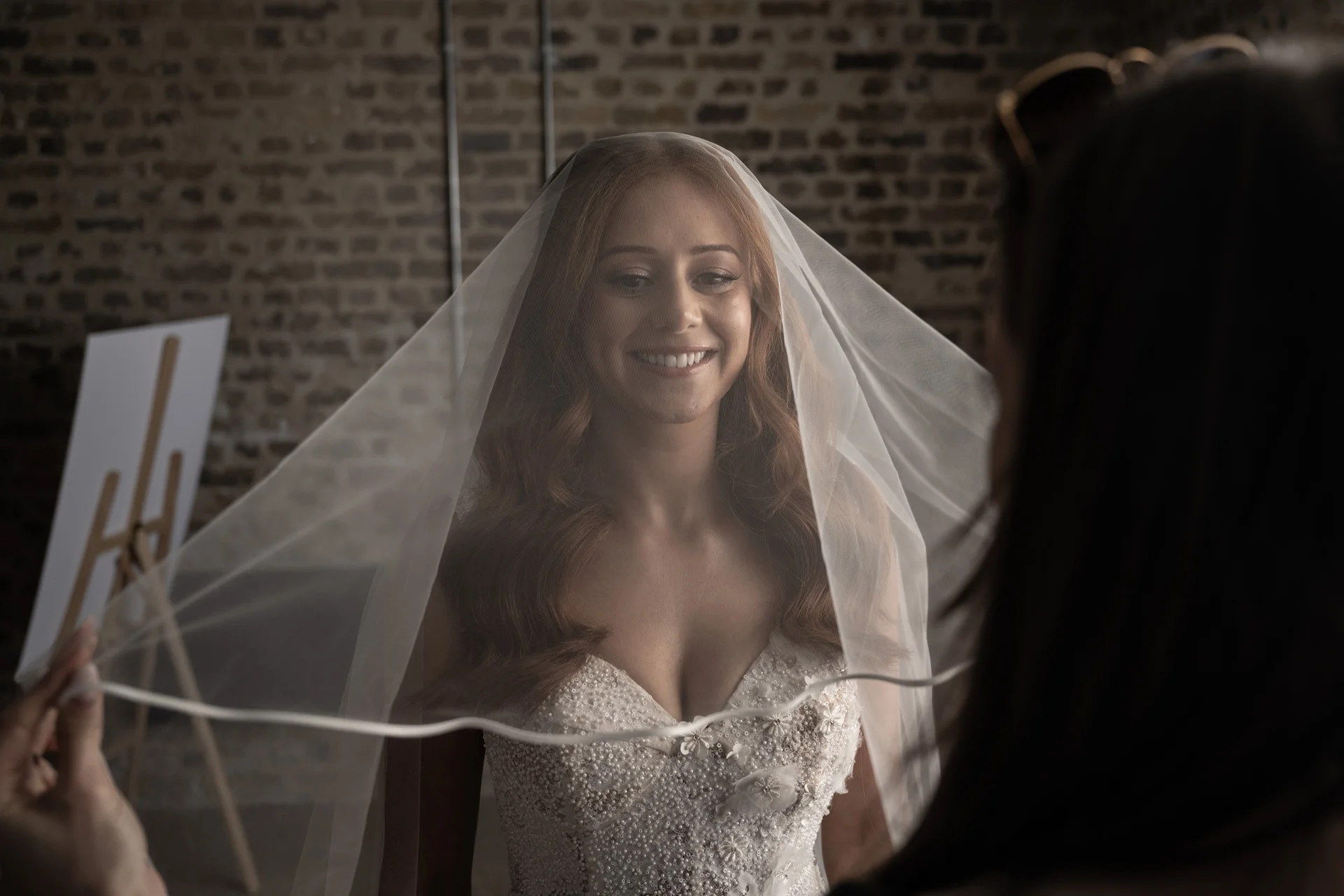 A smiling bride with red hair looks through a veil held by a stylist in front of an exposed brick wall during wedding preparations.