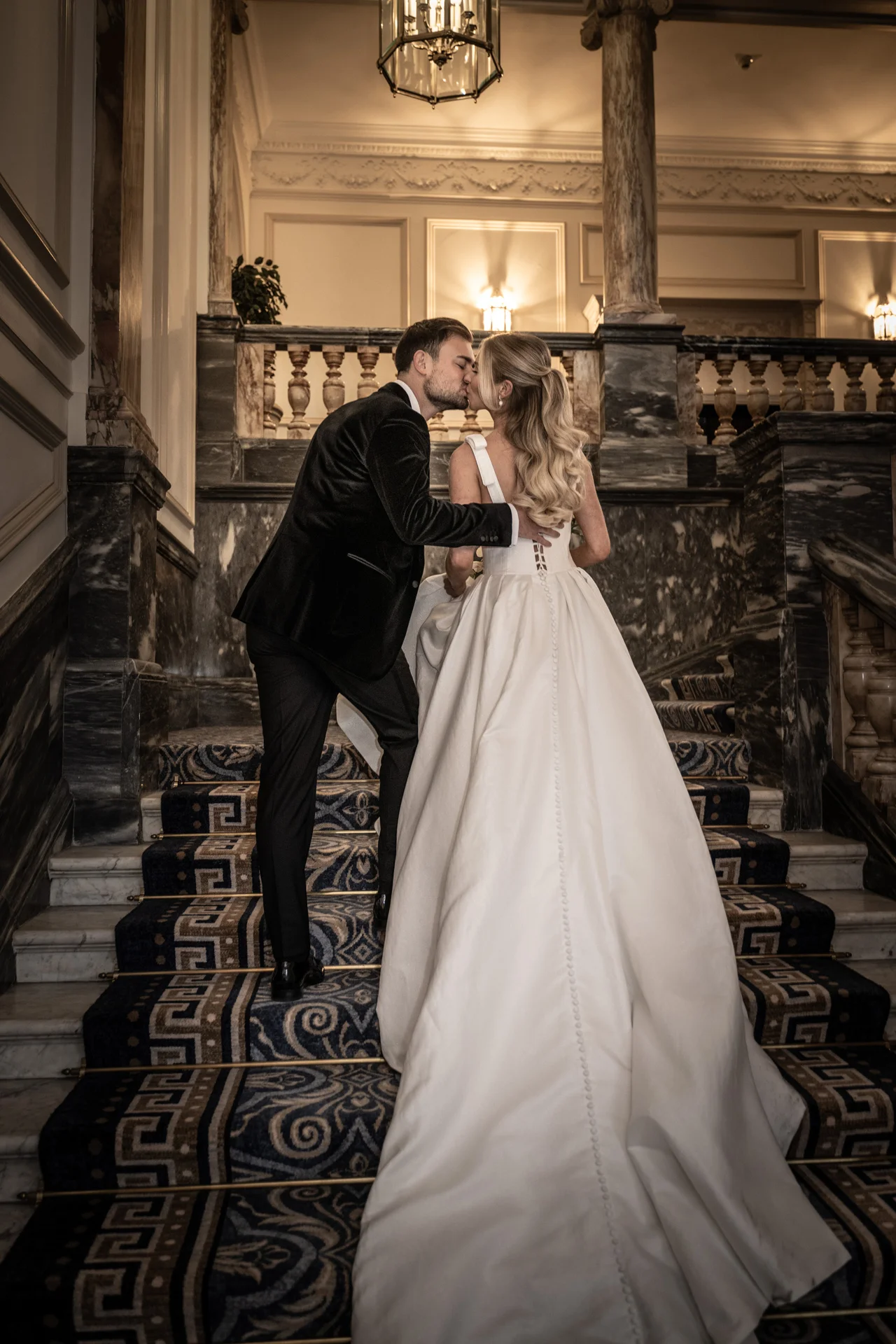A bride in an a-line satin gown and groom in a velvet tuxedo share a romantic kiss on a grand hotel staircase.