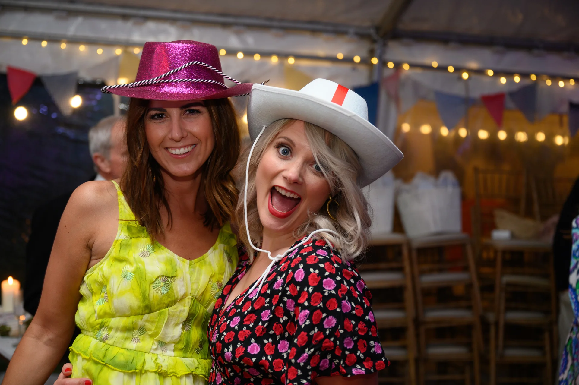 Two wedding guests wearing cowboy hats laugh joyfully inside a marquee lit with fairy lights and decorated with bunting.