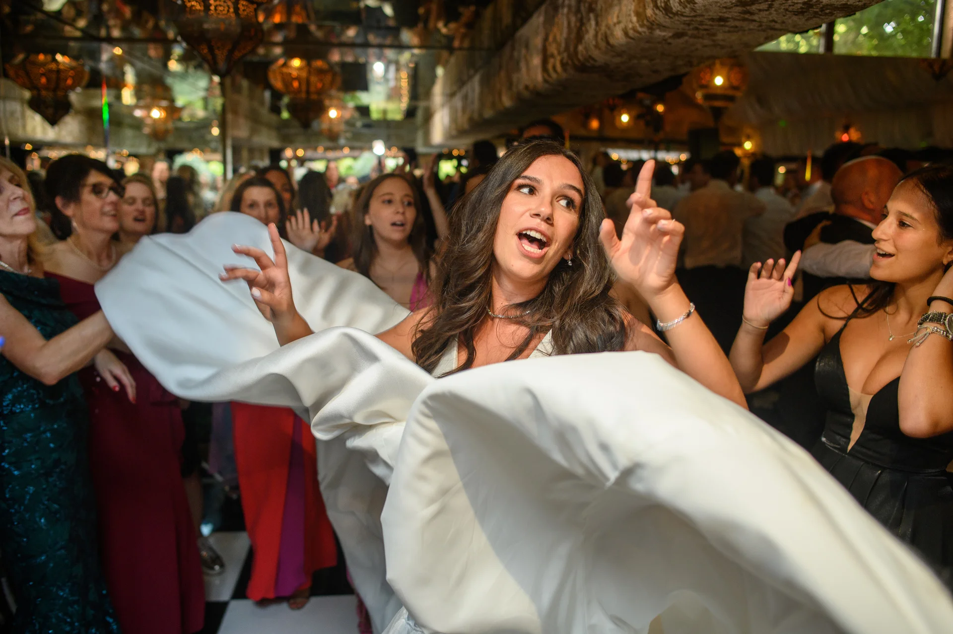 A joyful bride dances energetically on a checkered dance floor during the evening reception, her white satin dress swirling around her as guests cheer and dance in the background.