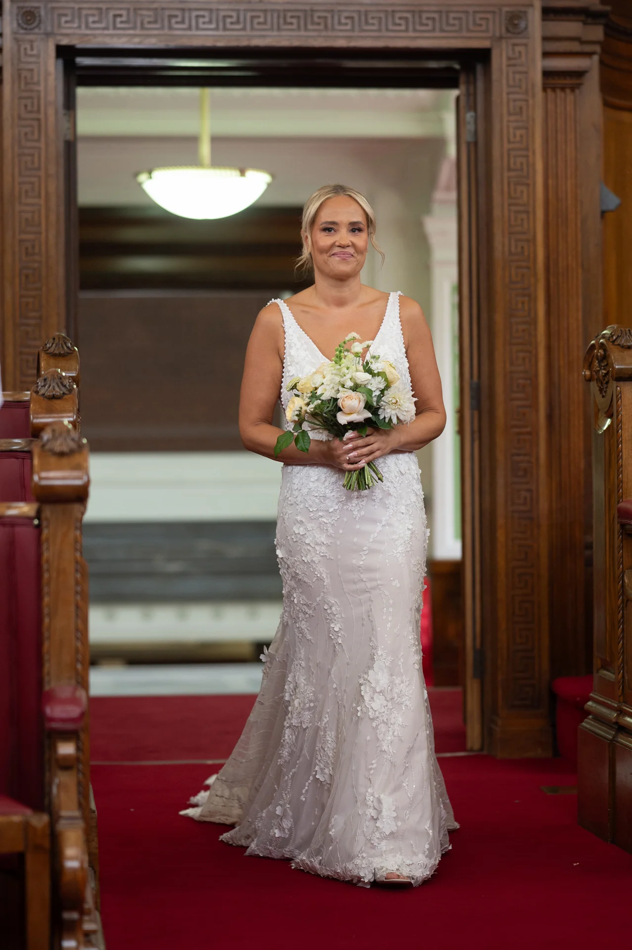 A smiling bride holding a hand-tied bouquet walks down the red-carpeted aisle of a traditional church, framed by an ornate wooden doorway.