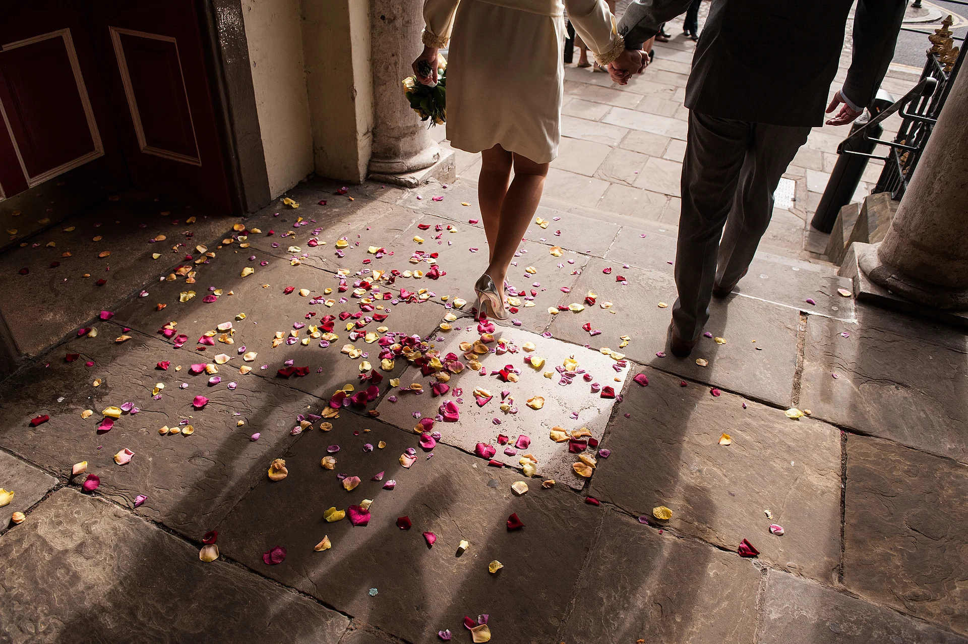 Newlywed couple walking away hand in hand across stone flagstones strewn with colourful confetti petals outside a historic building.