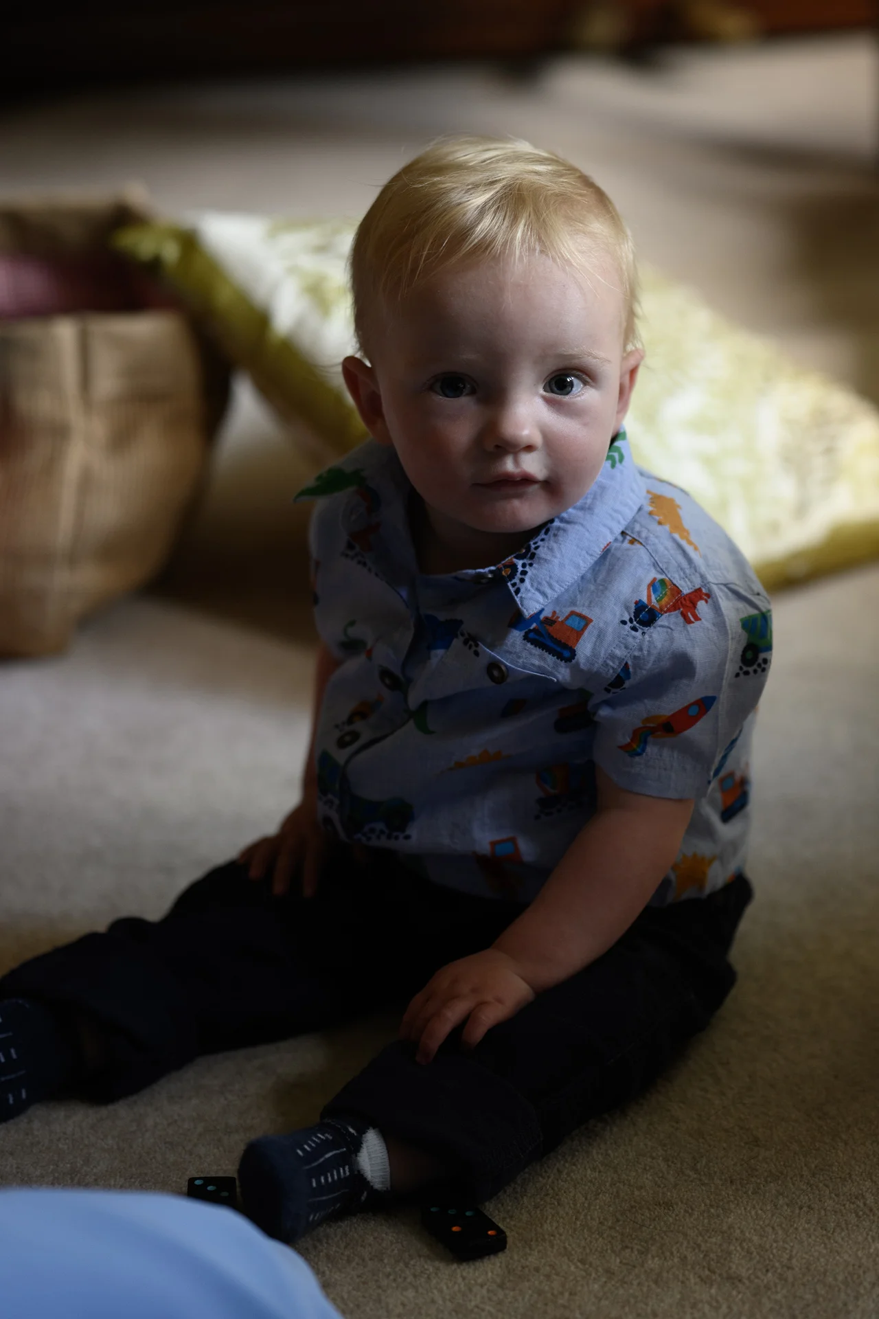 A young toddler with fair hair and a patterned blue shirt sits on the carpet playing with black dominoes during a wedding celebration.