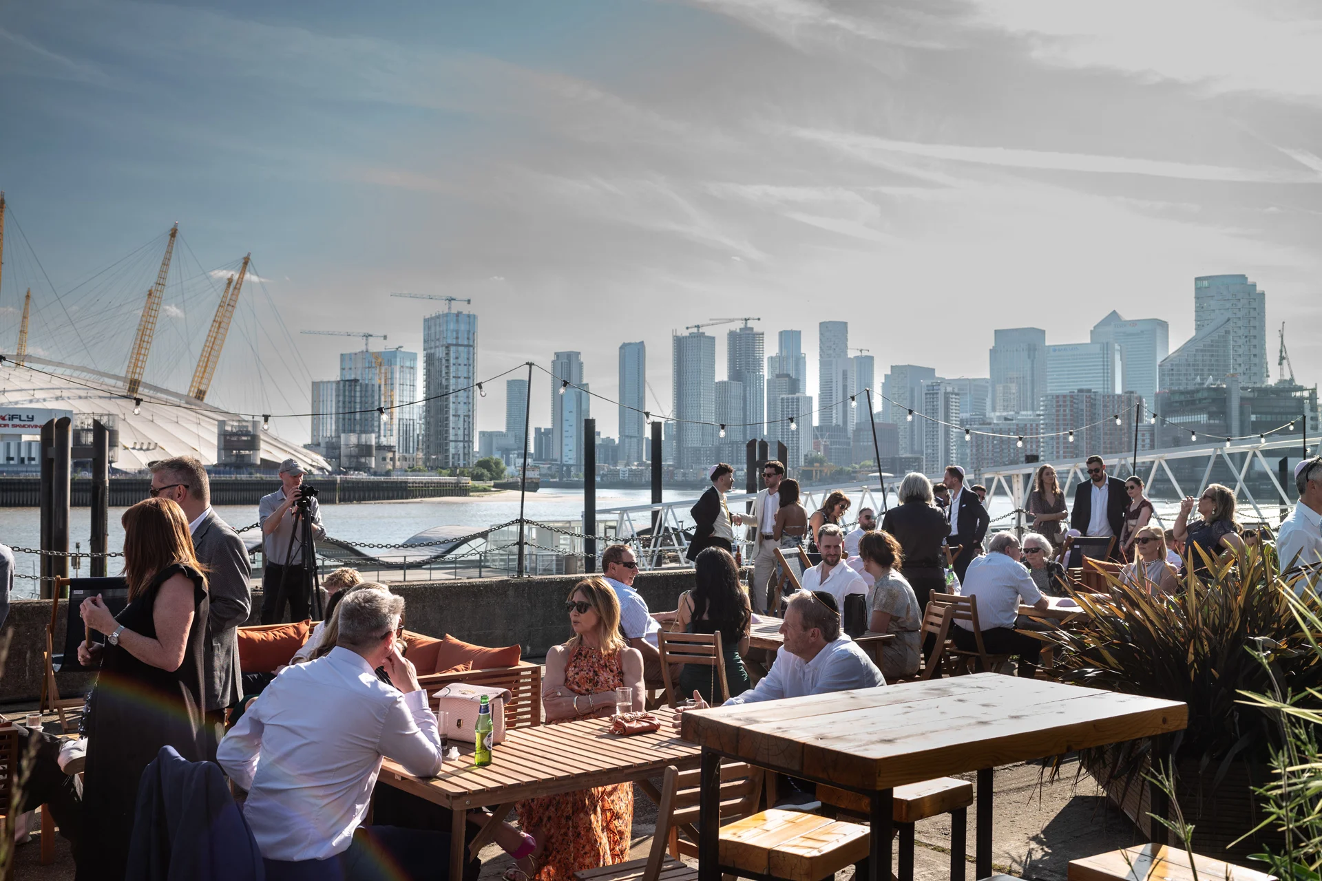 Wedding guests mingle during a sunny drinks reception on an urban rooftop terrace overlooking a city skyline and river.