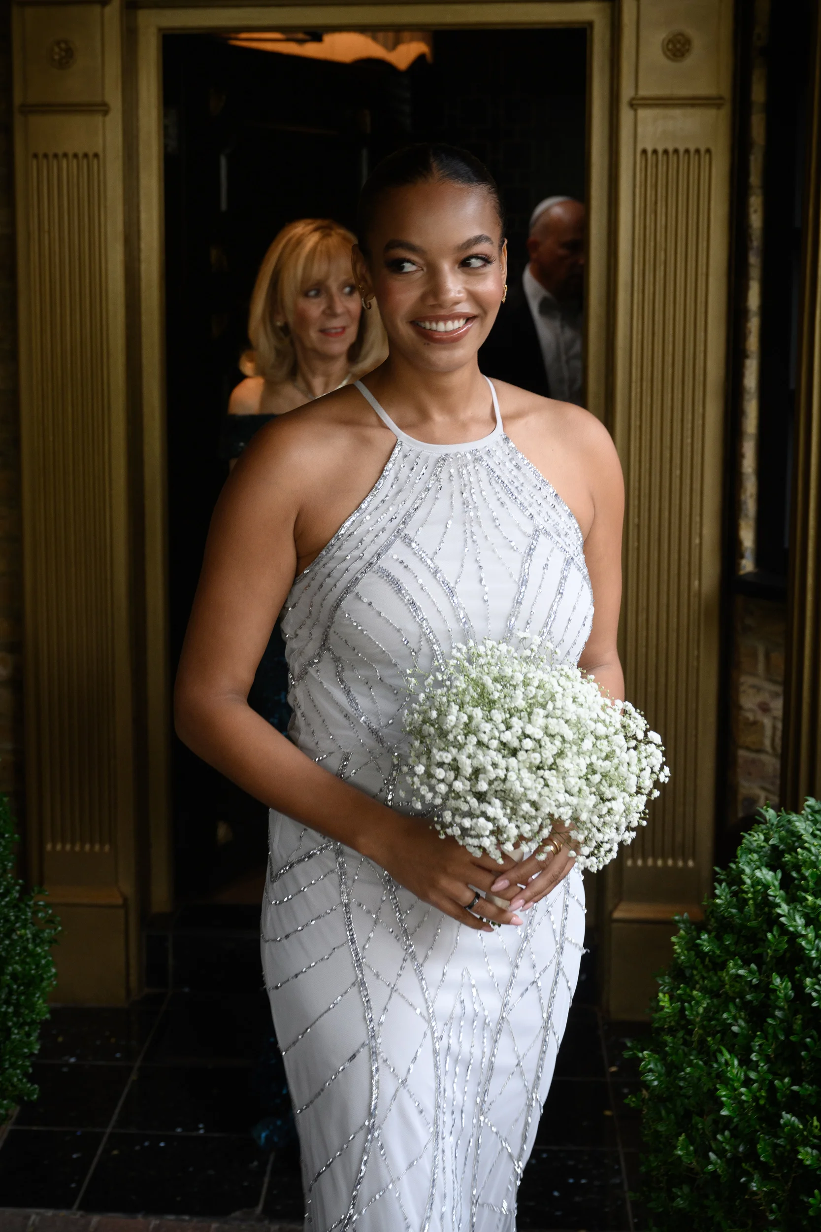 A smiling bride wearing an embellished halter-neck gown holds a baby's breath bouquet as she stands in a doorway.