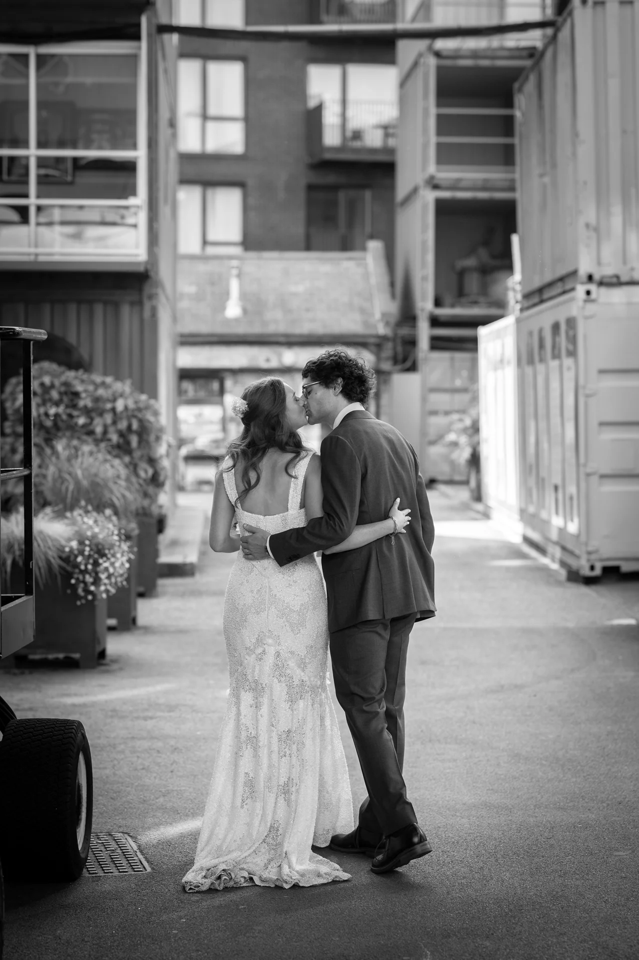 A newlywed couple shares a tender kiss while walking down an industrial city street flanked by shipping containers.