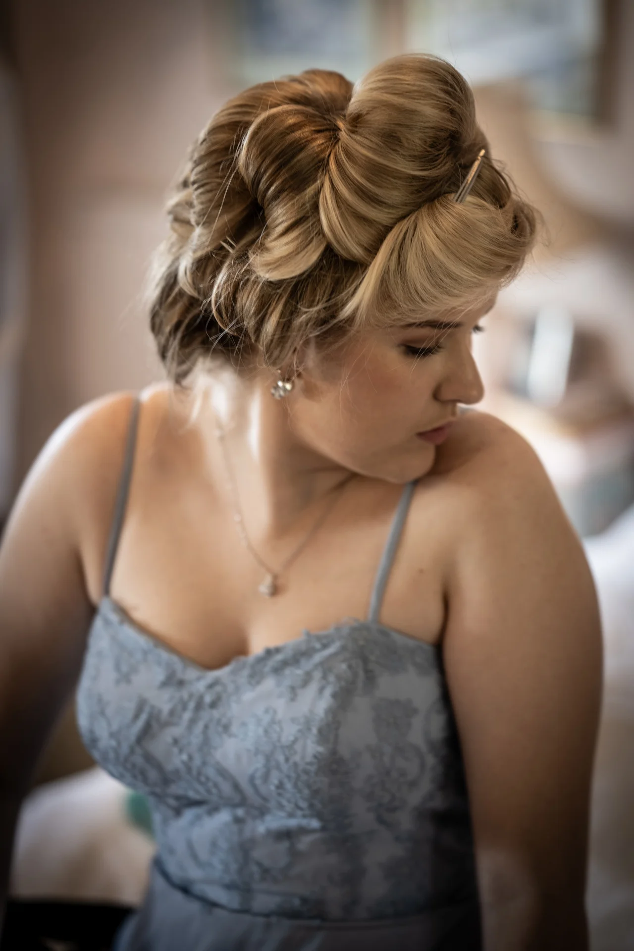 A close-up portrait of a blonde bridesmaid in a blue lace dress with a styled updo looking over her shoulder during morning preparations.