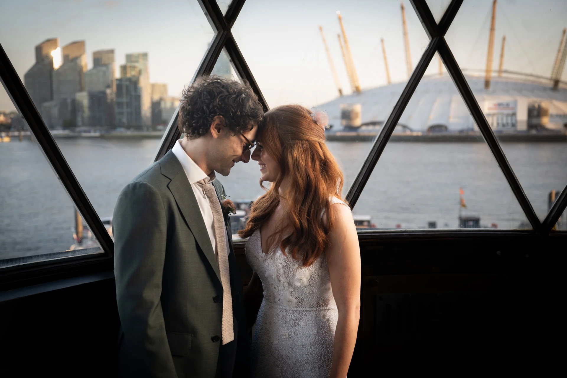 A newlywed couple stands nose to nose inside by a large geometric window, with a view of the London skyline and river visible in the background.