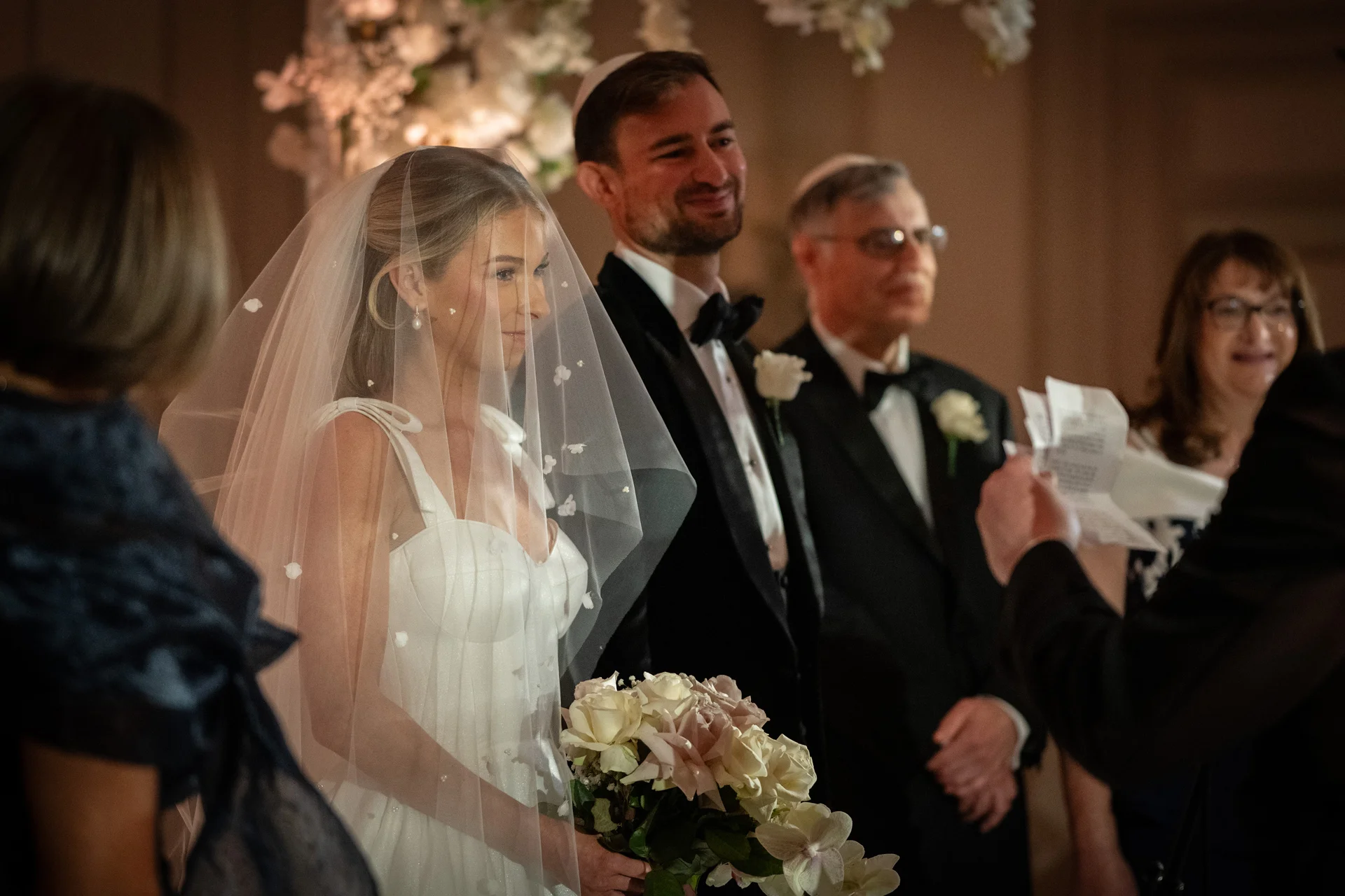A Jewish wedding ceremony captures the bride wearing a veil and holding a bouquet alongside the groom in a tuxedo, with guests and family watching.
