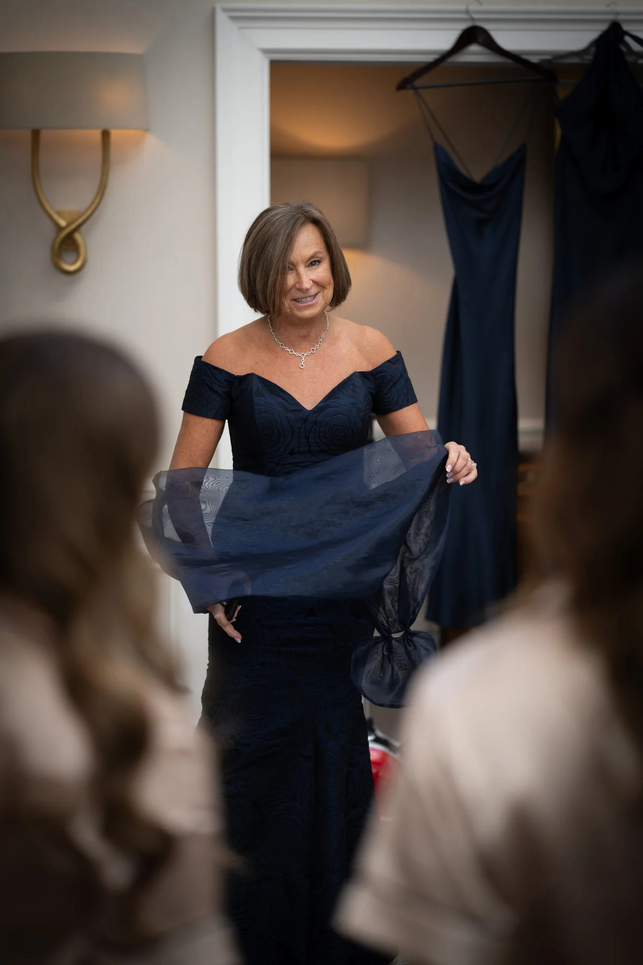 The mother of the bride smiles while trying on a dark navy evening gown in a brightly lit dressing room, framed by guests in the foreground.
