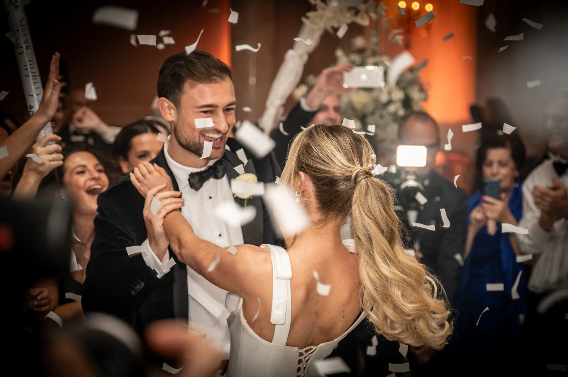 Newlyweds share their first dance surrounded by cheering guests as white confetti falls around them in celebration.