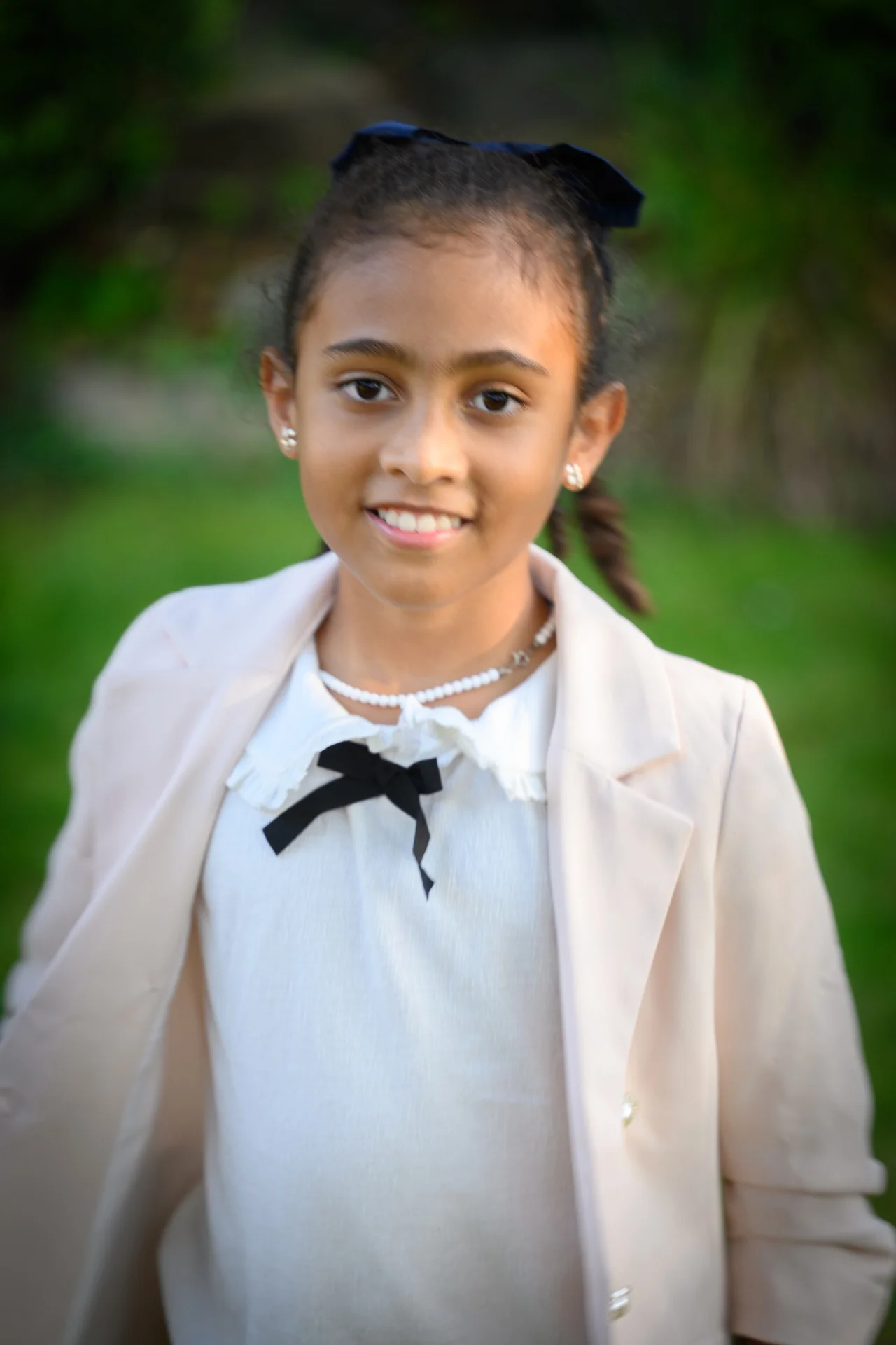 A young flower girl smiles outdoors against a soft green garden background, wearing a light-coloured blazer over a white top adorned with a black bow.