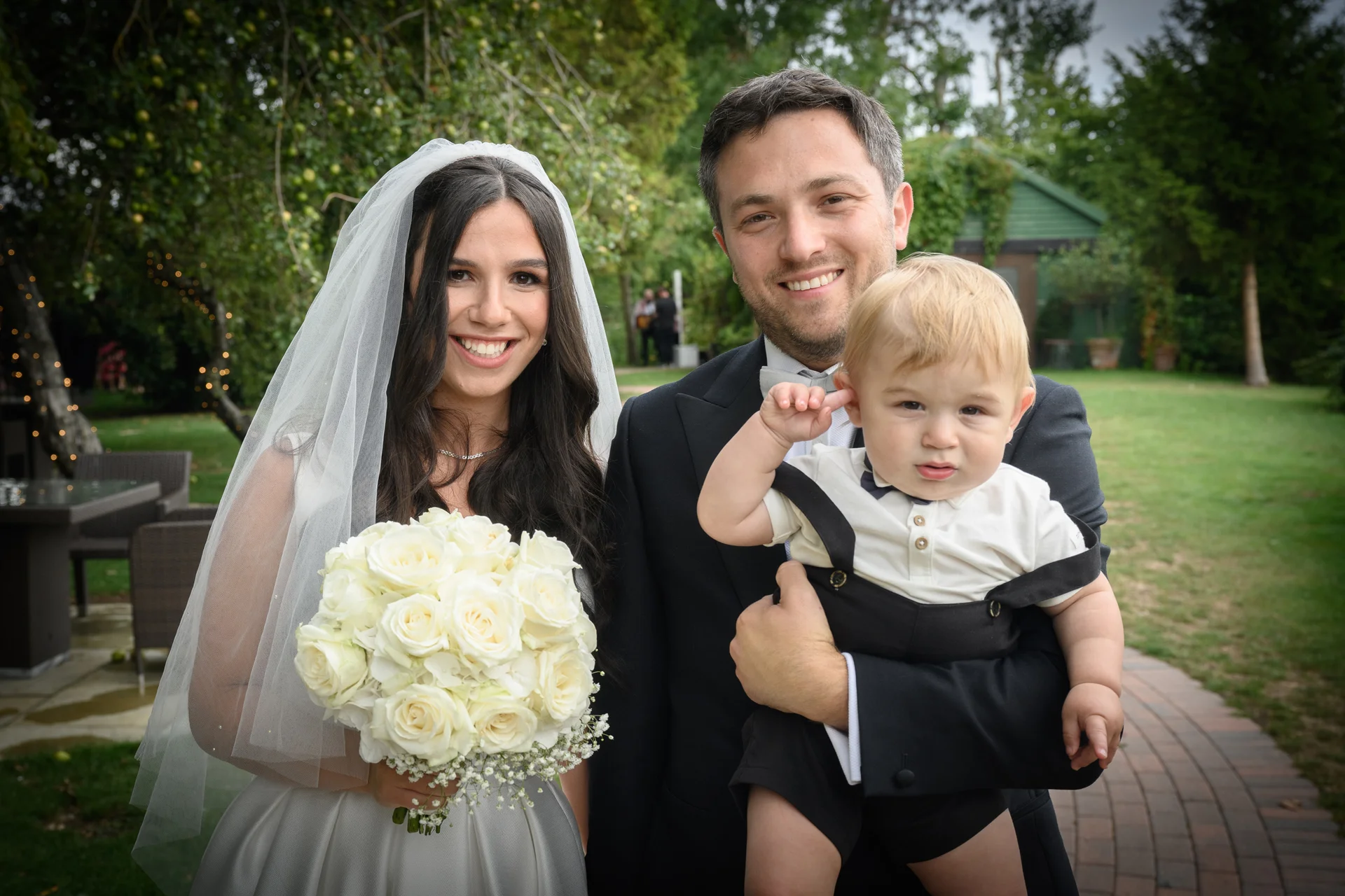 A smiling bride holding a white rose bouquet and groom holding a toddler page boy pose together outdoors in a lush garden setting.