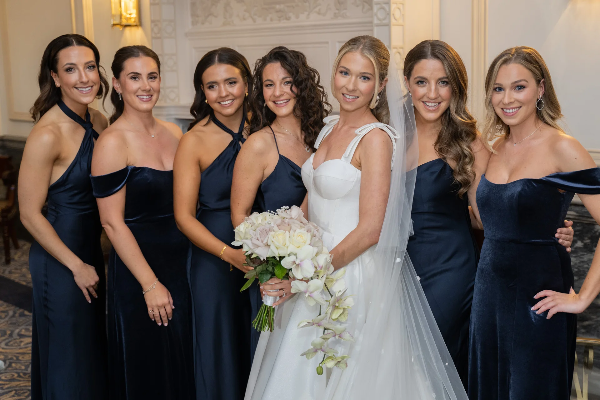 A smiling bride holding a cascading bouquet stands alongside her bridesmaids dressed in chic navy gowns.