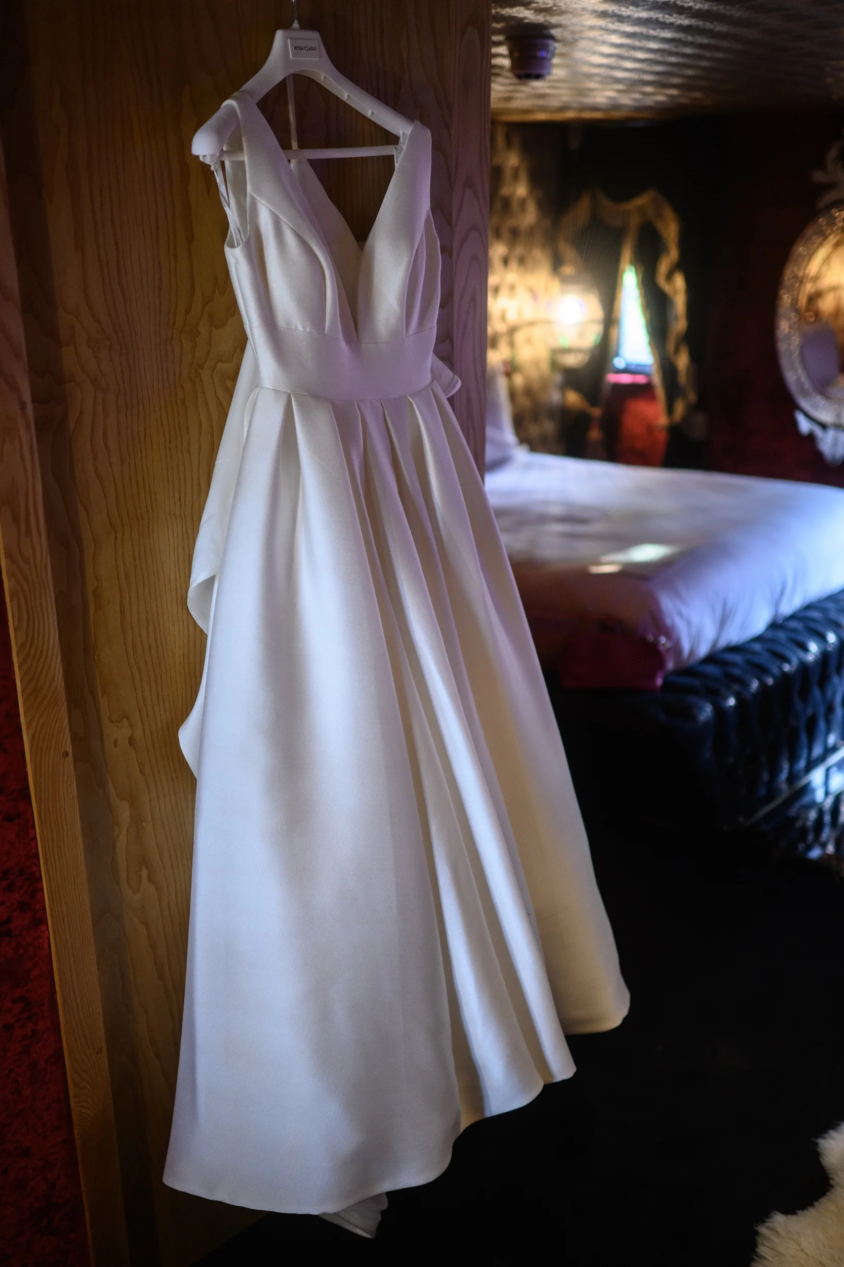 An ivory a-line wedding gown with a plunging v-neckline and pleated skirt hangs neatly from a wooden panel inside a warmly lit bridal preparation room.