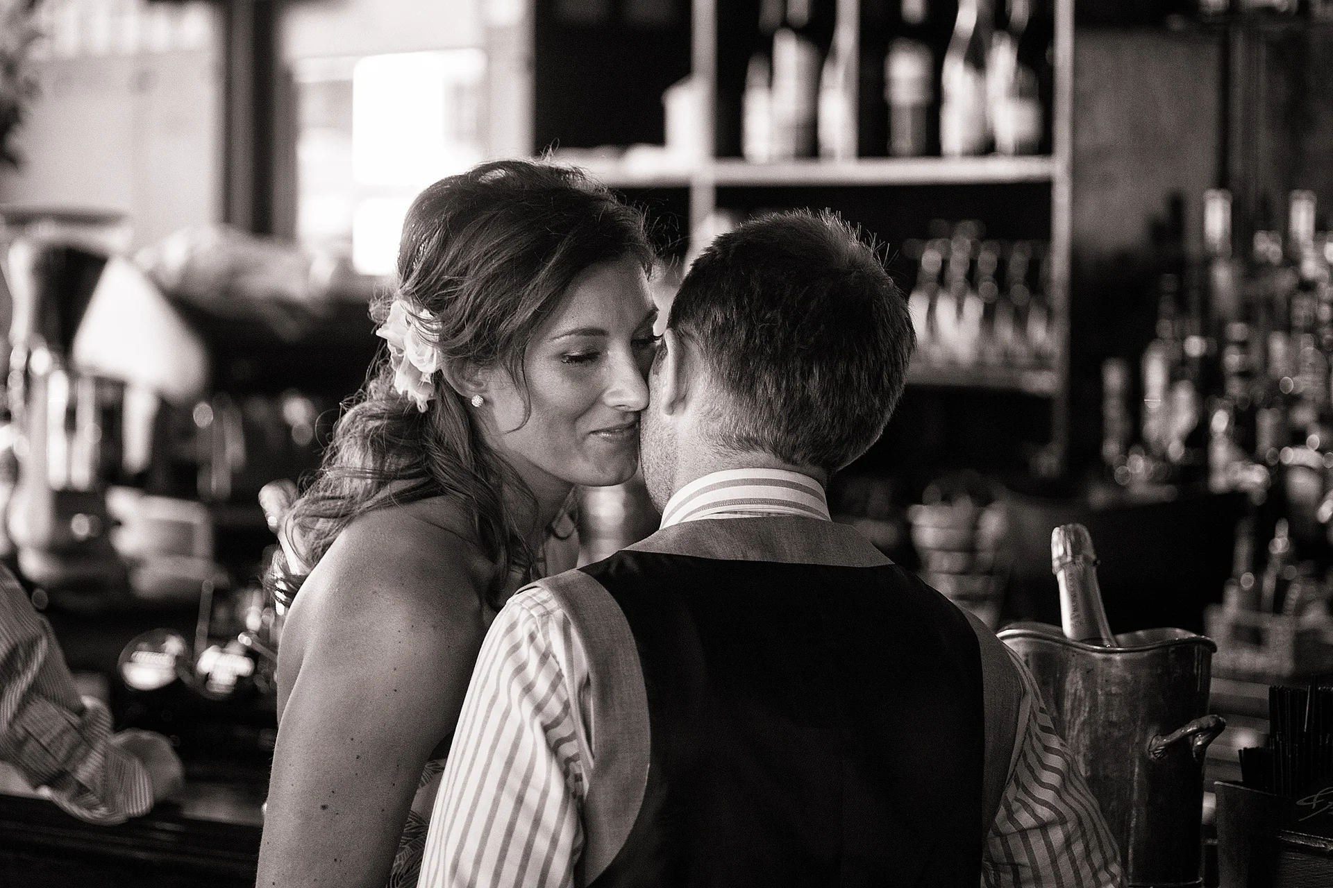 A newlywed couple shares a tender whispered moment across a rustic pub bar in a striking black and white photograph.