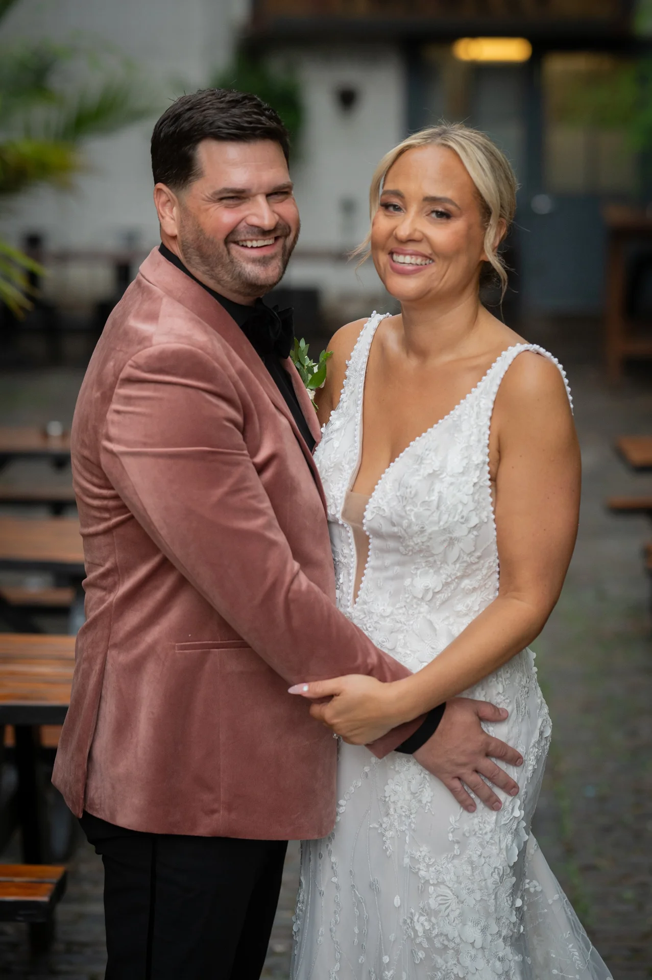 A smiling bride in a plunge neck lace wedding dress and a groom in a pink velvet tuxedo jacket pose together outdoors.
