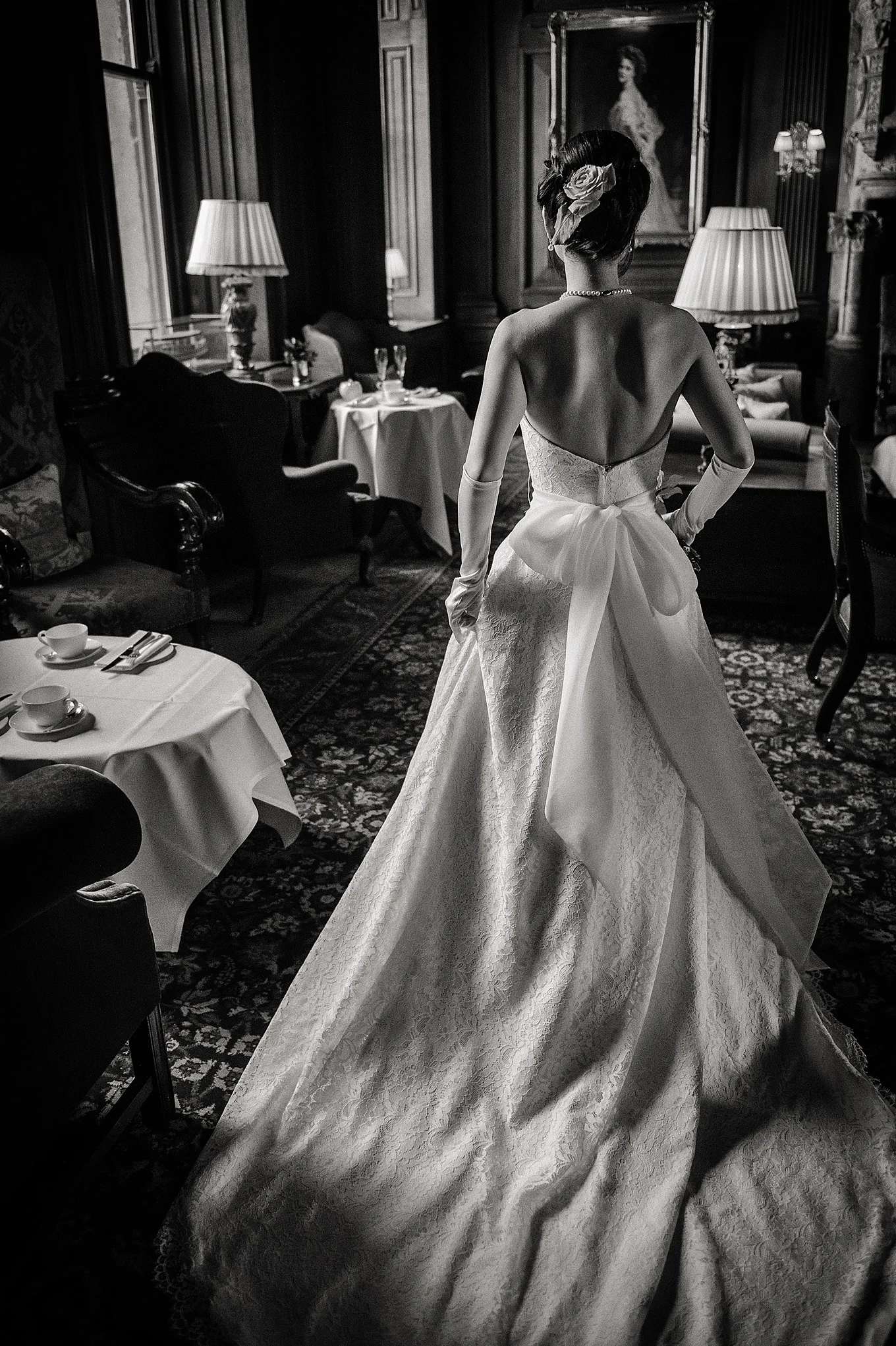 A stunning black and white portrait captures a bride from behind inside an elegant stately home, showcasing her lace wedding gown with a dramatic train and oversized bow.