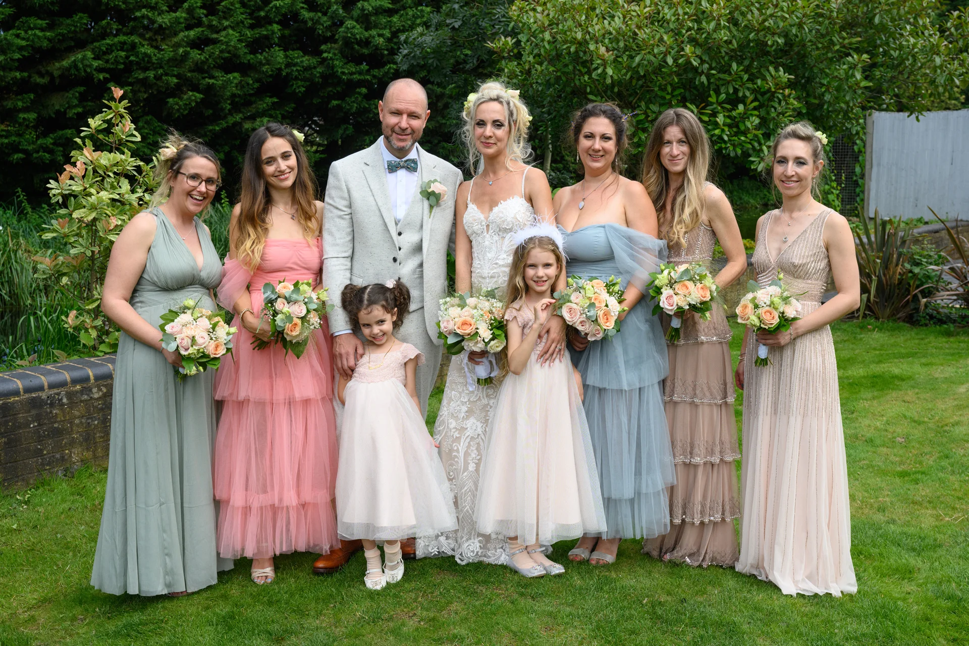 A smiling wedding party poses outdoors in a lush garden setting, featuring a bride in a lace gown, a groom in a tweed suit, and bridesmaids in mismatched pastel dresses holding floral bouquets.