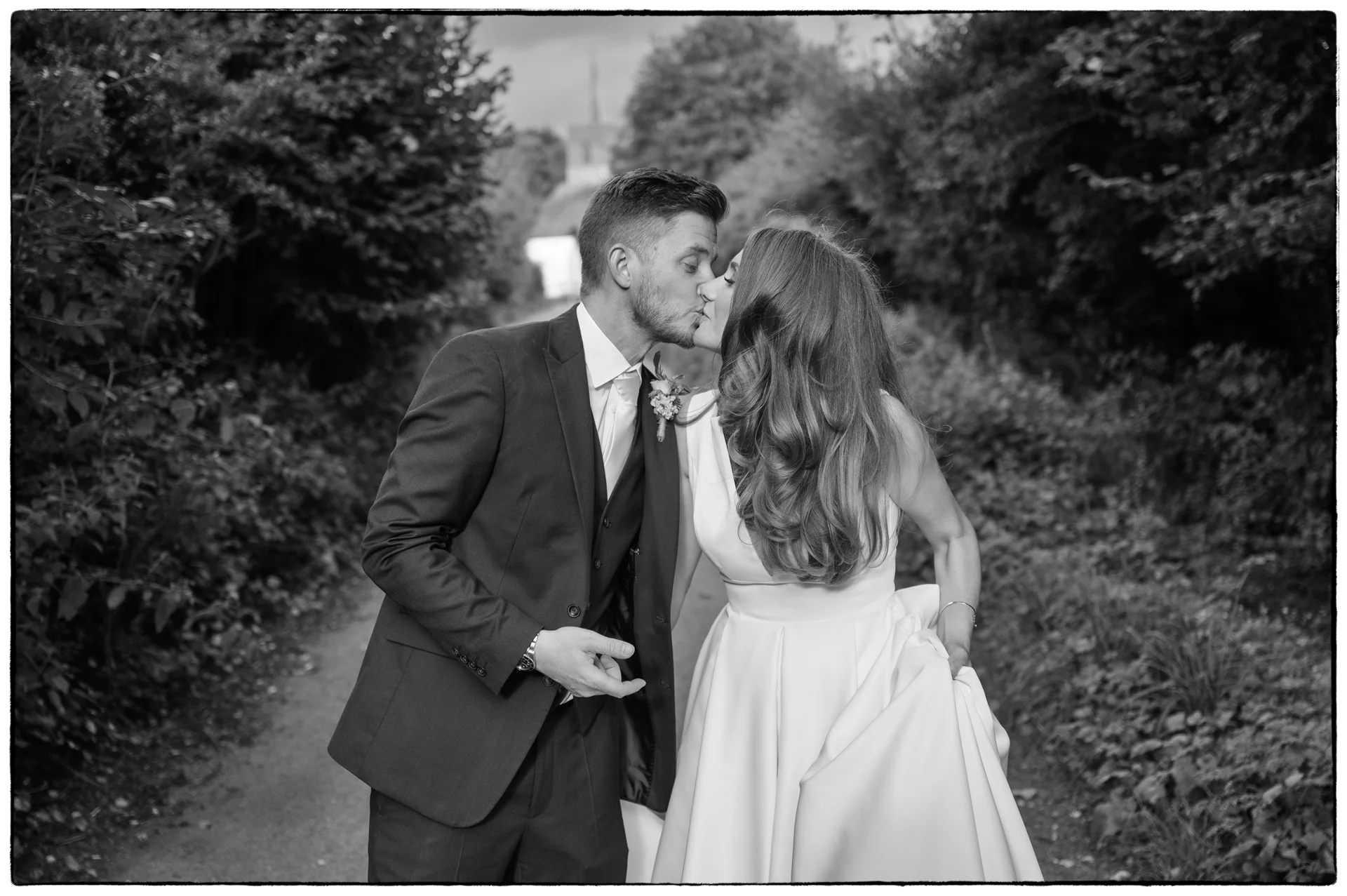 Newlyweds share a tender kiss along a scenic, tree-lined path in this classic black and white portrait.