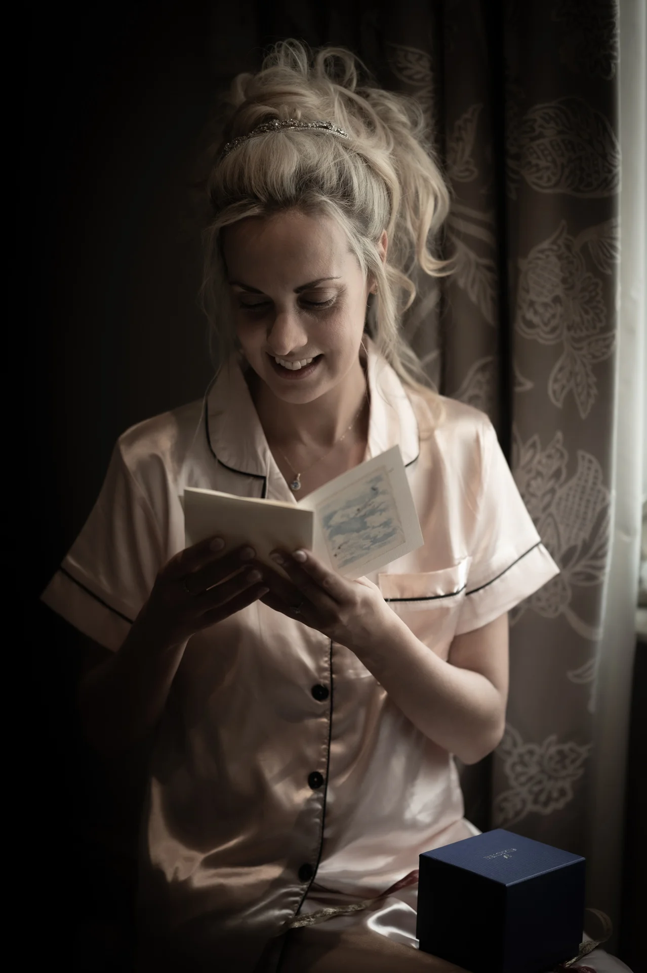 A smiling bride wearing satin pyjamas sits by a window while reading a heartfelt letter during morning preparations.