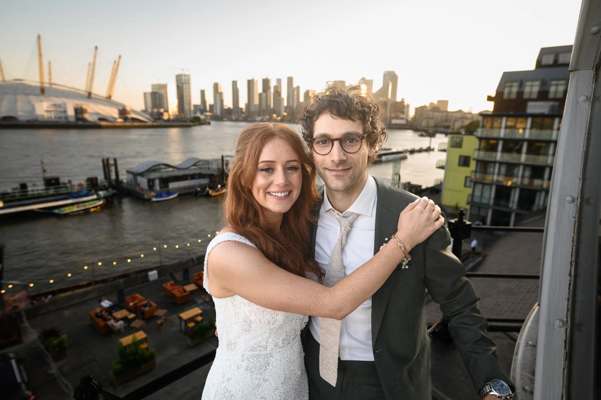 A newlywed bride with red hair in a lace dress and a groom in a green suit pose together on a rooftop overlooking the London skyline at sunset.