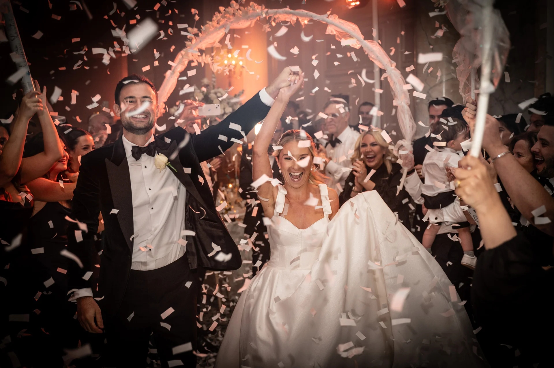 A joyful bride and groom walk through a cheering crowd amidst a shower of white confetti during their evening reception entrance.