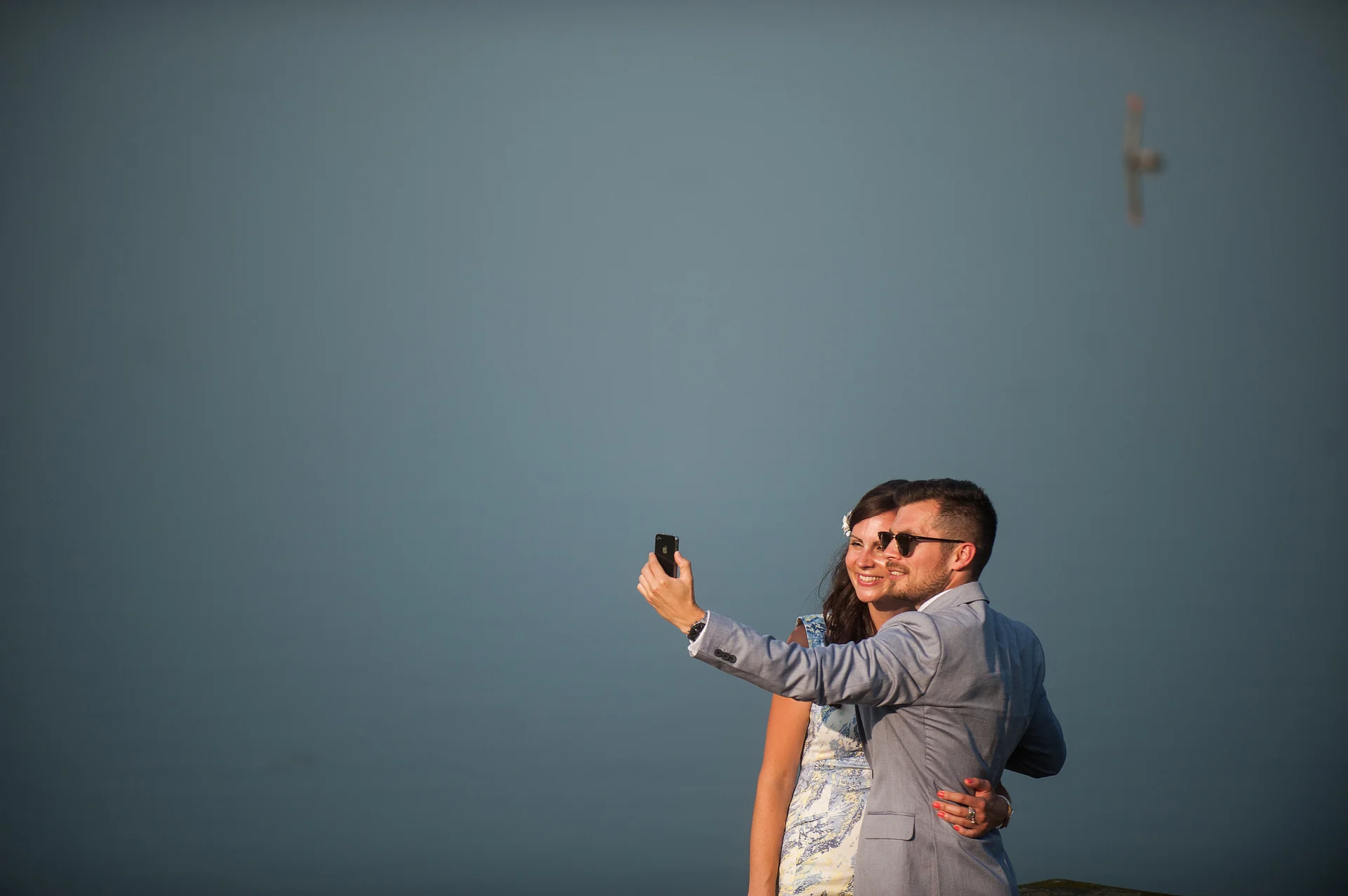 A newlywed couple in a light-coloured suit and patterned dress take a selfie together outdoors against a muted background.
