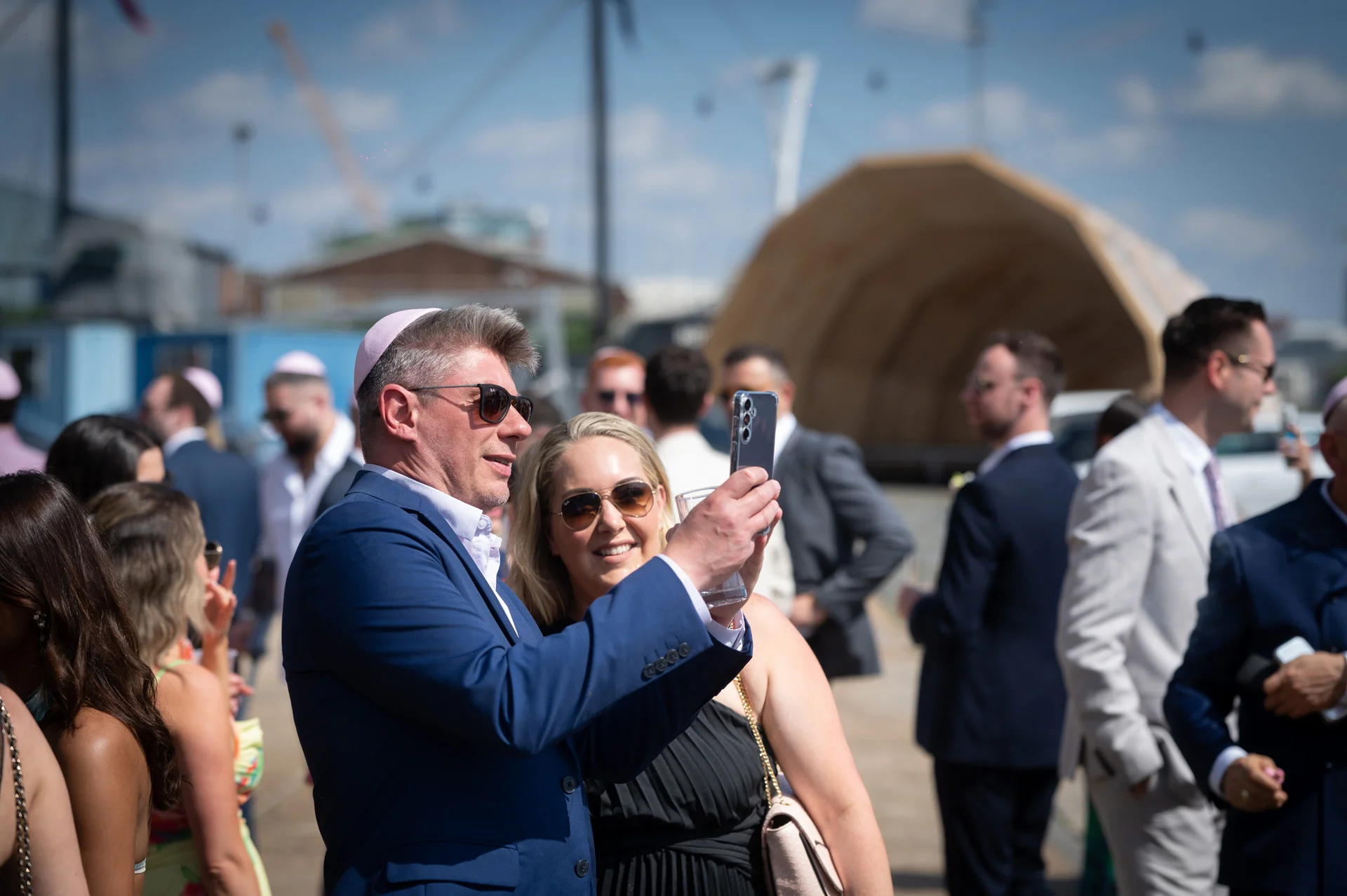 Wedding guests in sunglasses and formal attire take a selfie together in the bright outdoor sunshine during the daytime celebration.