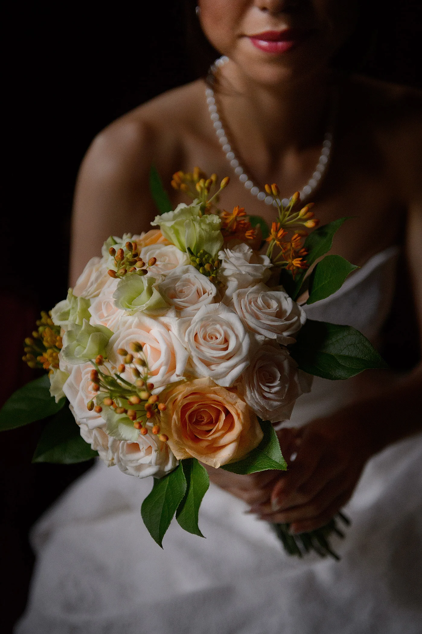 A close-up portrait of a bride holding a round hand-tied bouquet of peach and cream roses against a dark backdrop.