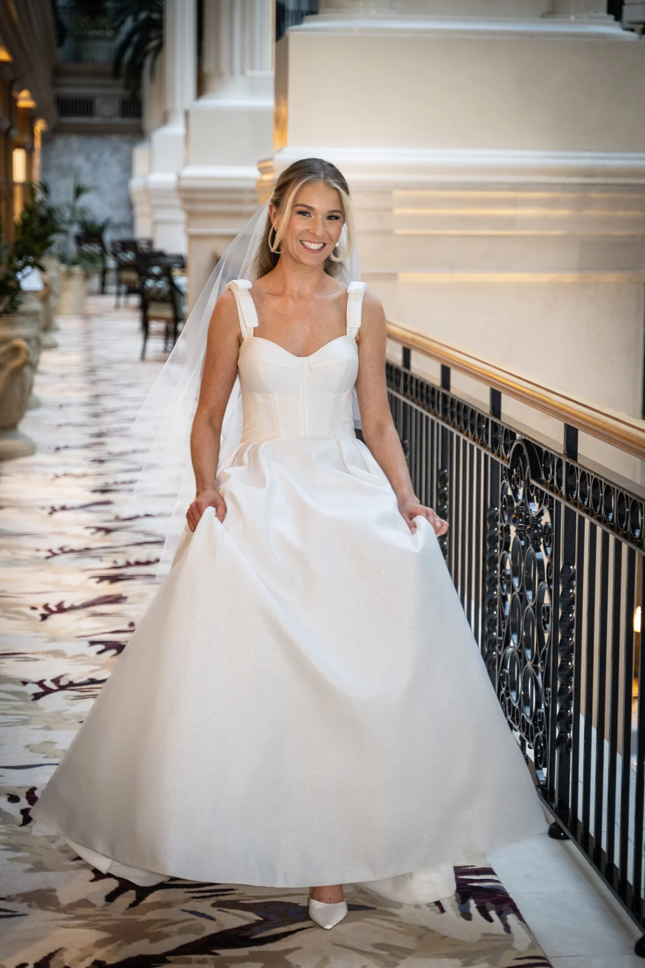 A smiling bride wearing an A-line satin gown with a sweetheart neckline and a veil poses on a hotel landing next to a staircase.