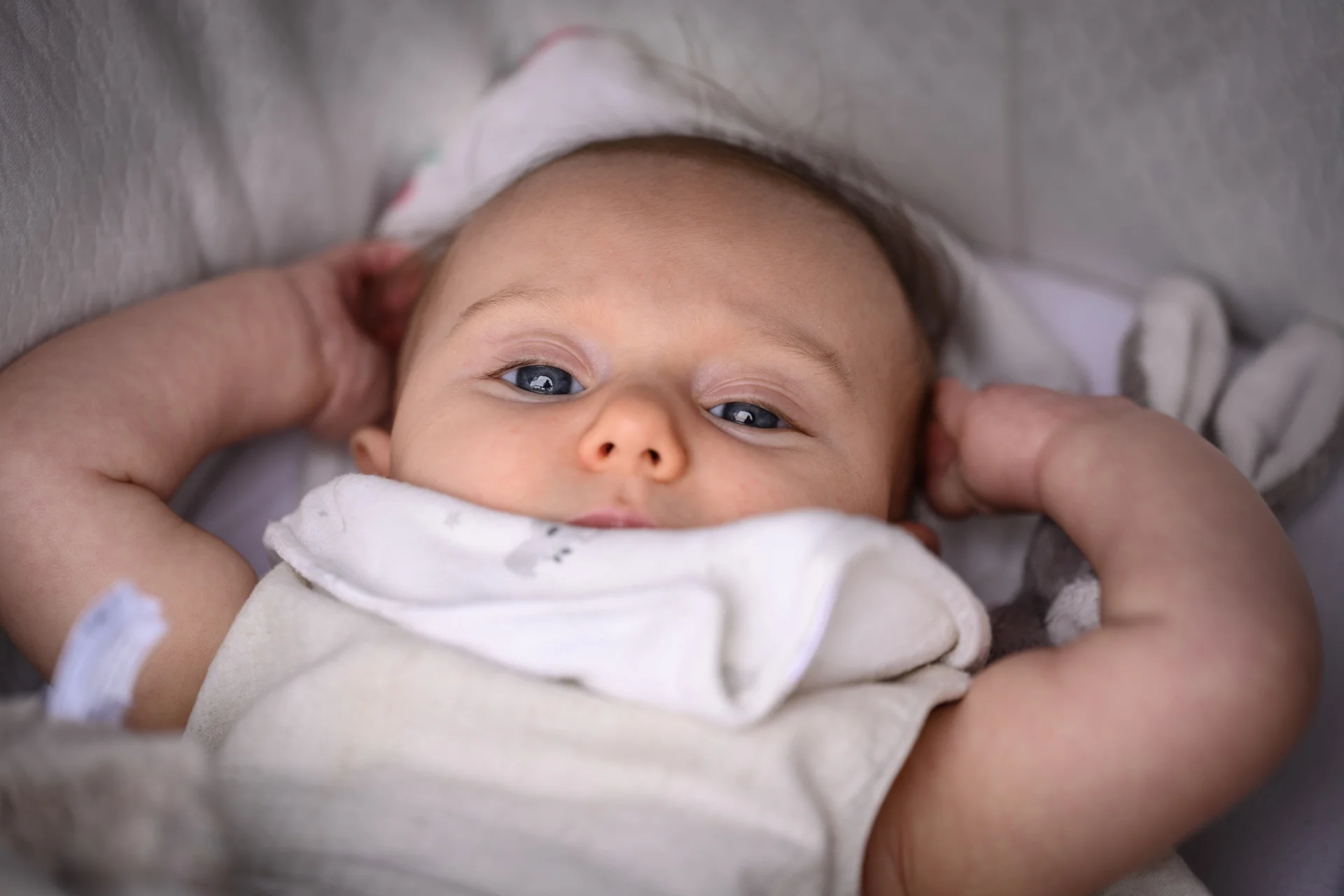 A close-up photograph of a baby lying down with hands up near its head, captured in soft natural light.