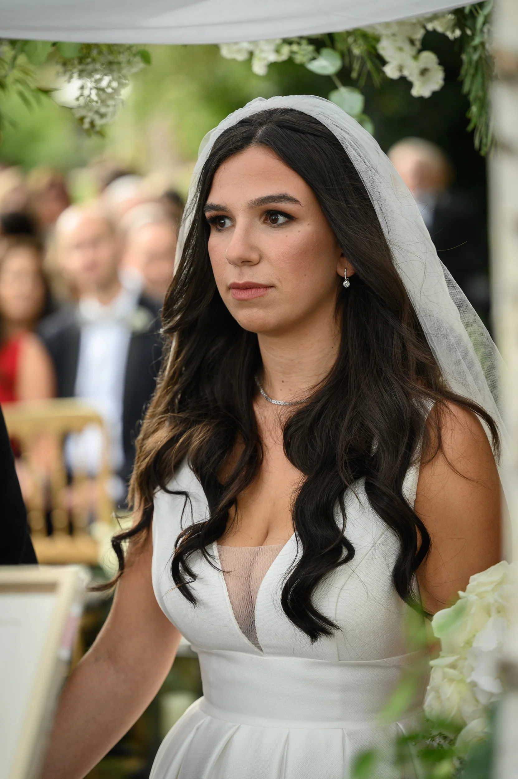 A stunning dark-haired bride wearing a white V-neck gown and a delicate veil stands at an outdoor wedding ceremony.