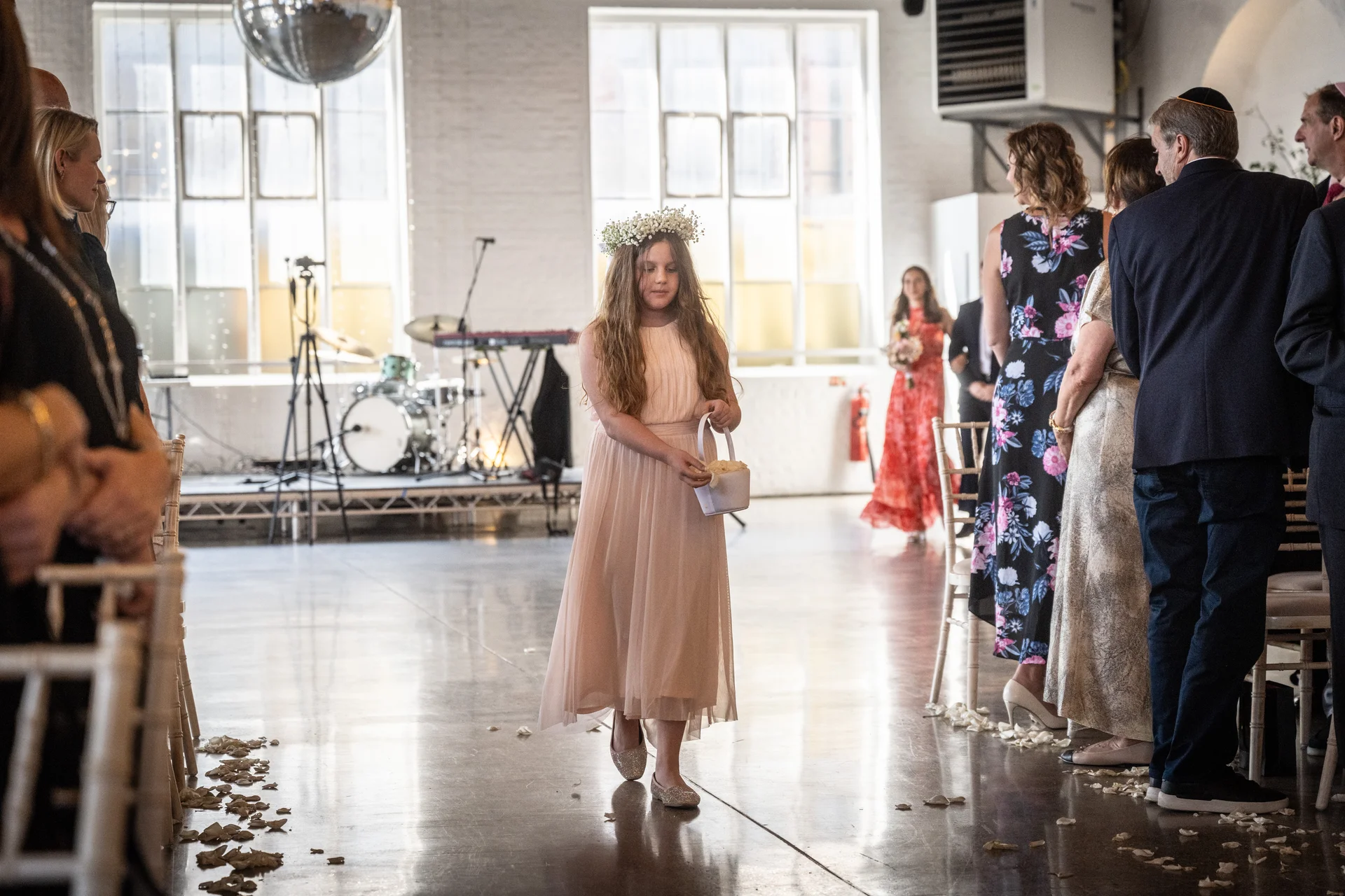 A young flower girl in a pink dress and floral crown walks down the aisle during a ceremony in an industrial warehouse venue.