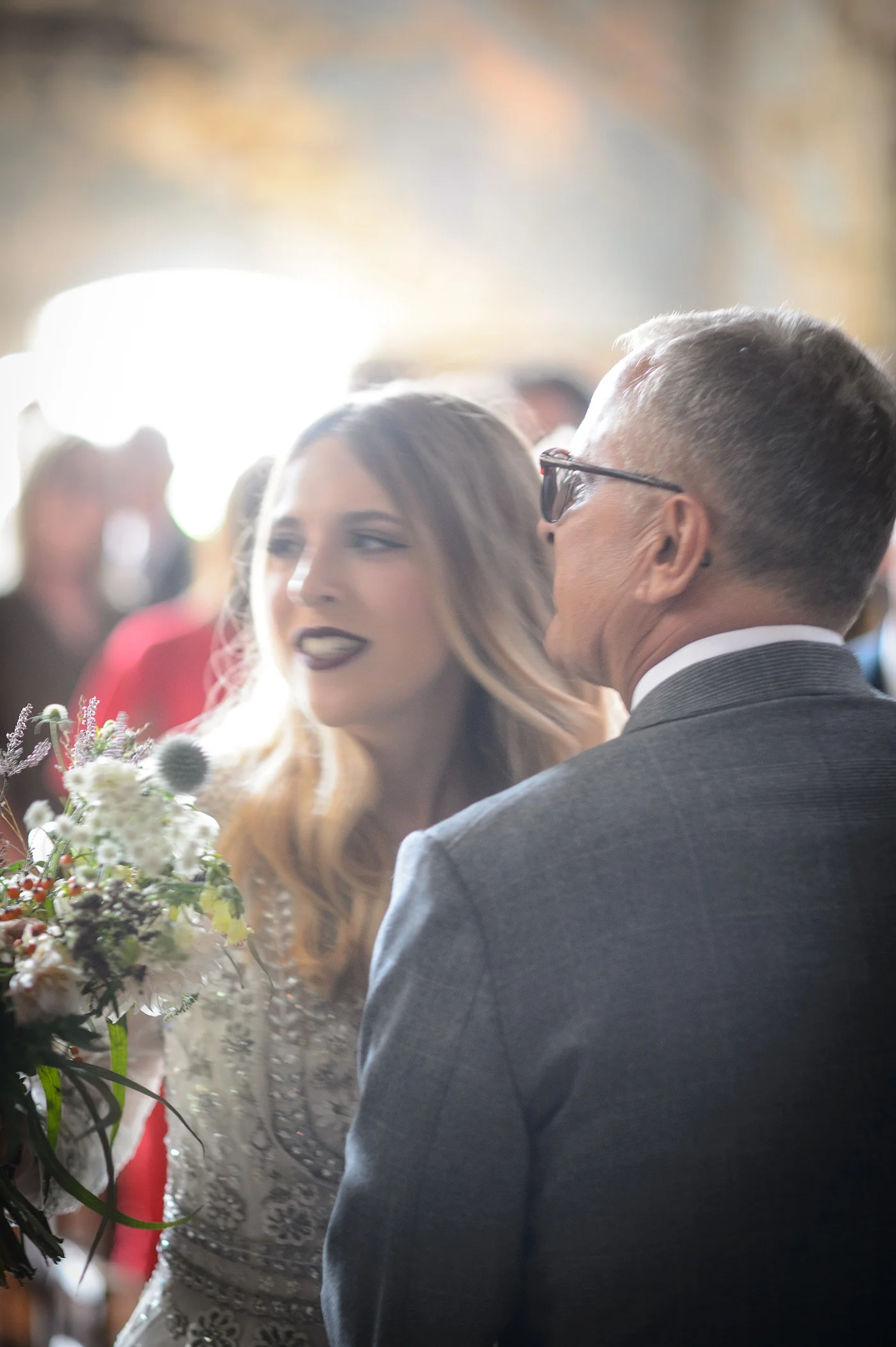 A smiling bride holding a textured bouquet shares a tender moment with the father of the bride wearing a grey suit.