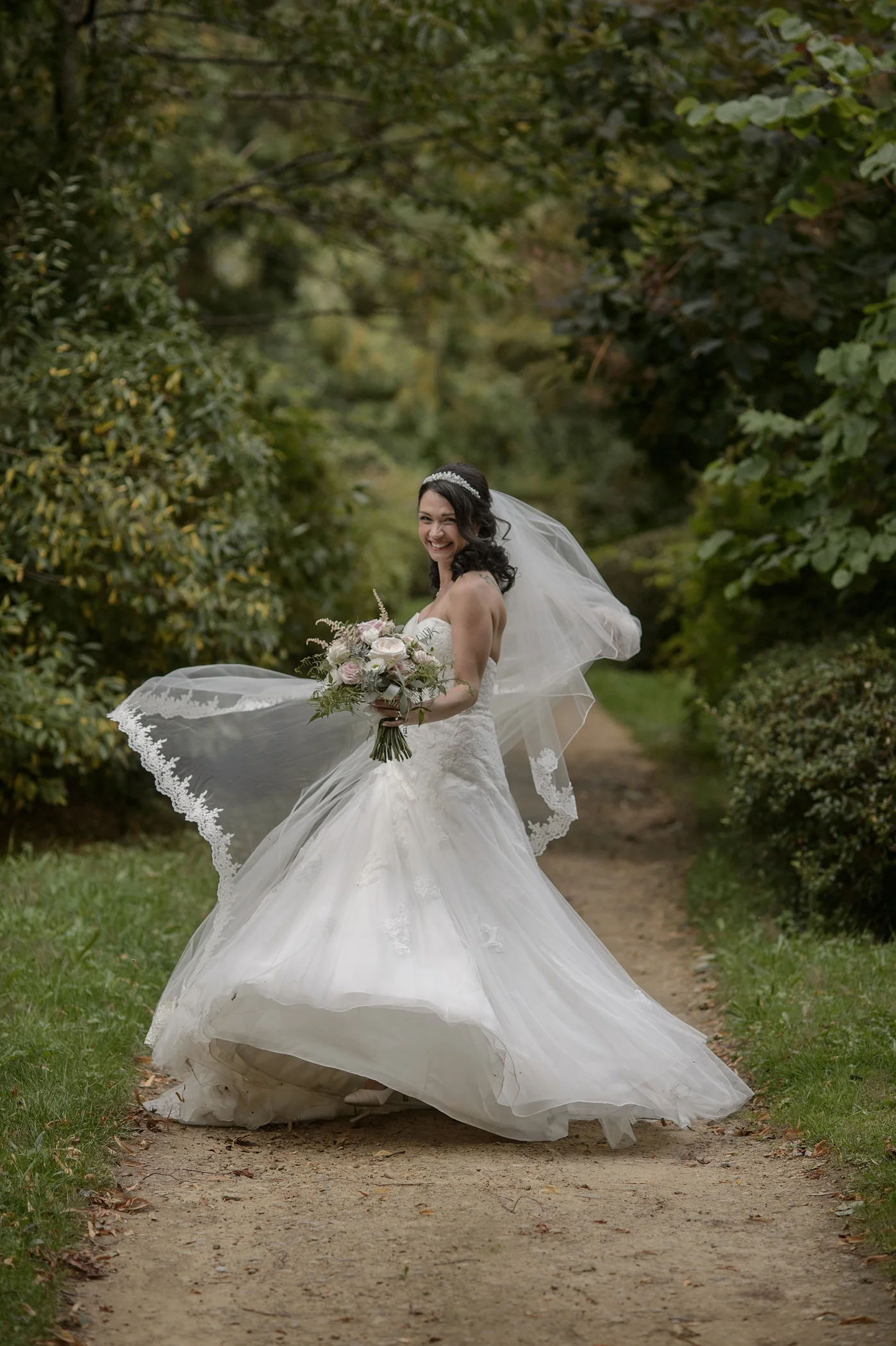 A smiling bride wearing a strapless lace and tulle wedding dress twirls on a woodland path, her veil billowing in the wind as she holds a hand-tied bouquet.