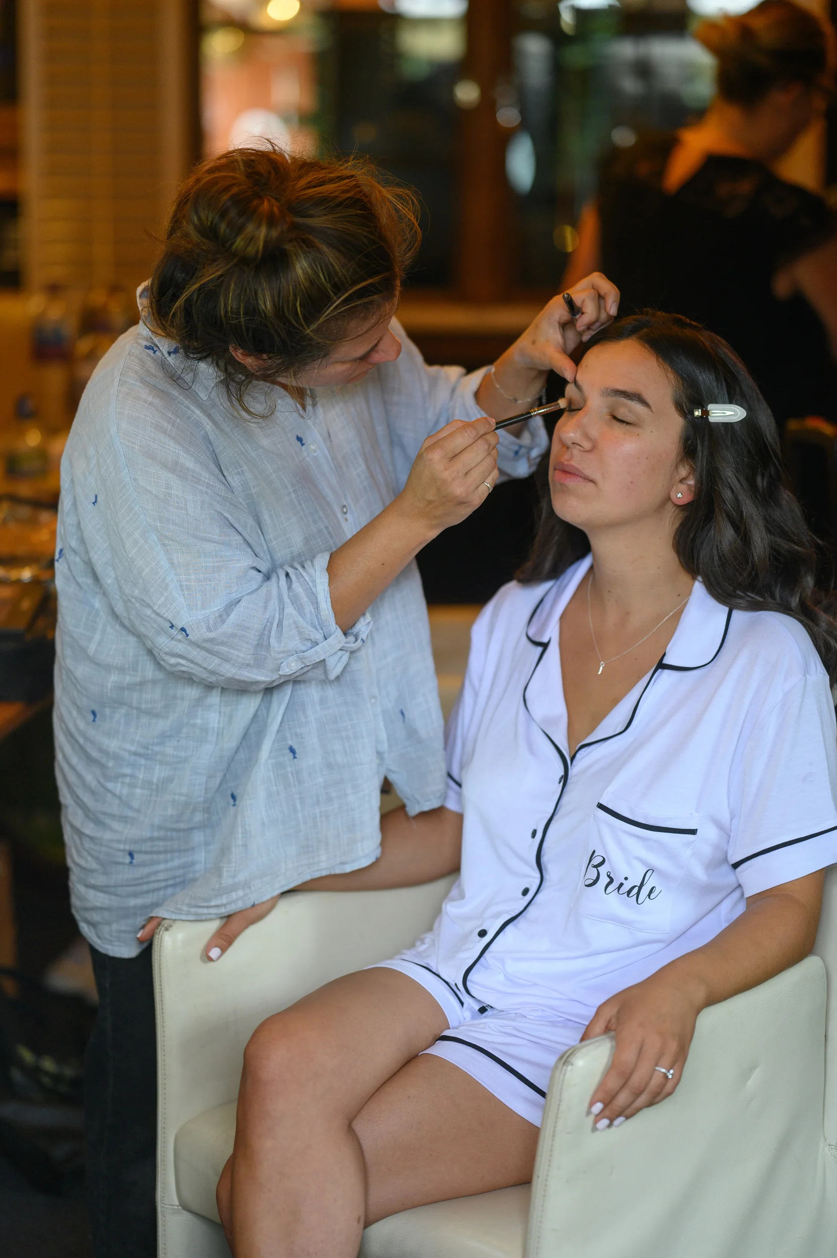 A professional makeup artist applies eye makeup to a bride who is seated and wearing personalised matching pyjamas during morning bridal preparations.