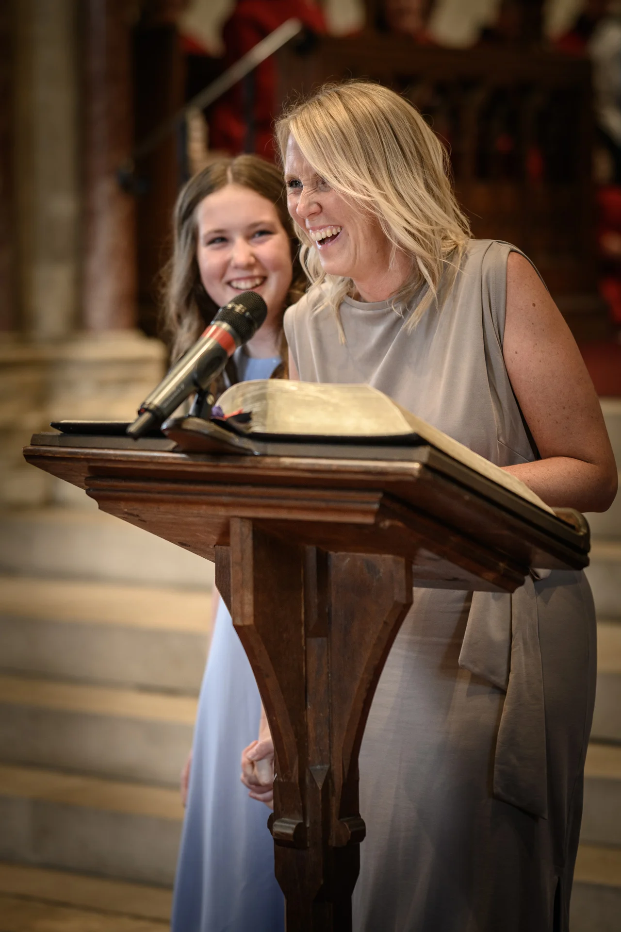 A smiling woman reading from a book at a wooden lectern during a church wedding ceremony with another guest standing nearby.