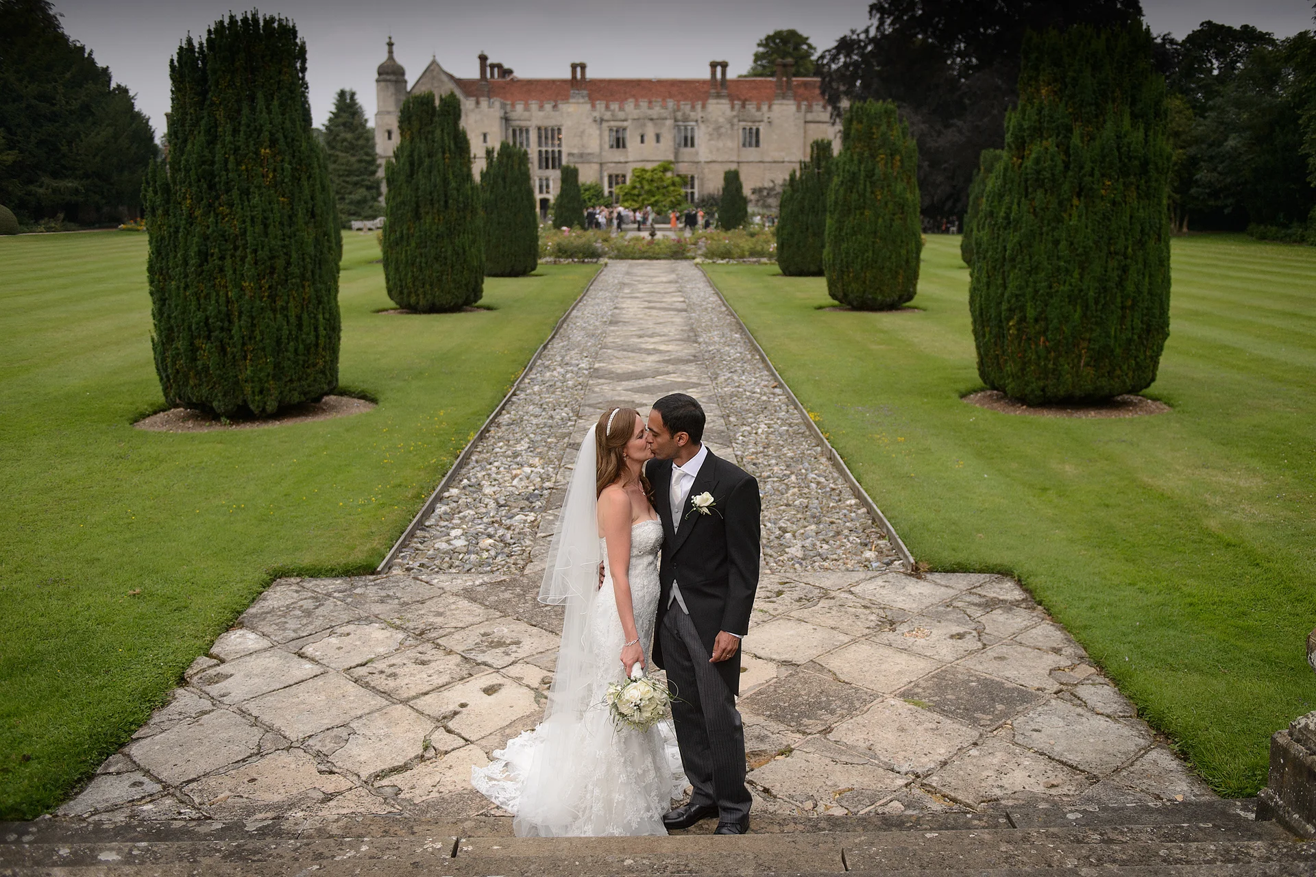 A newlywed couple shares a kiss on a stone terrace in front of a grand manor house with manicured lawns and towering topiary trees.