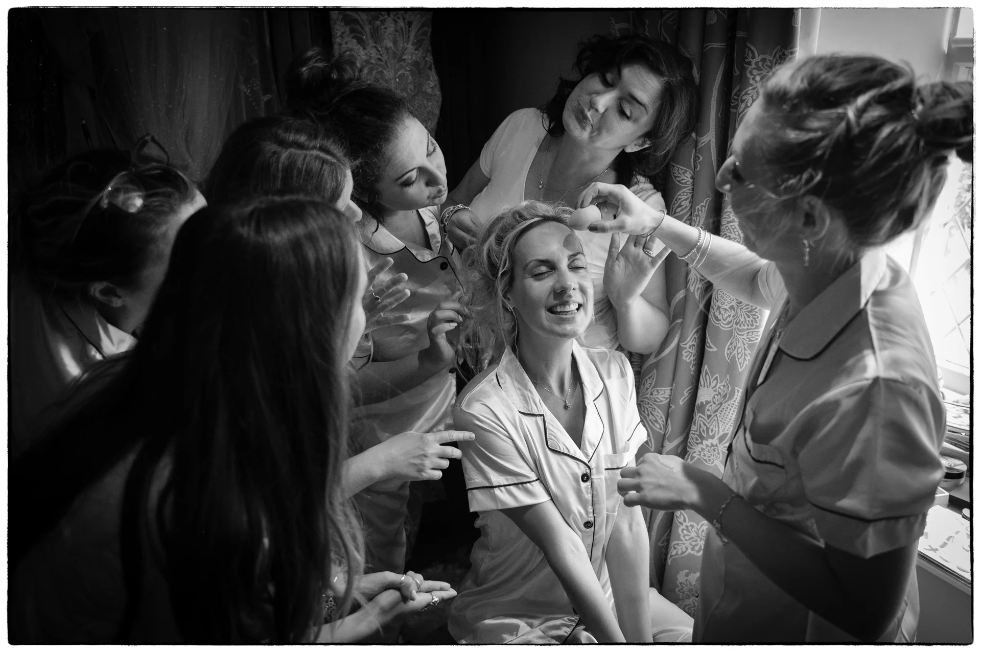 A laughing bride sits surrounded by her bridesmaids in matching pyjamas as she gets her makeup applied during morning bridal preparations.