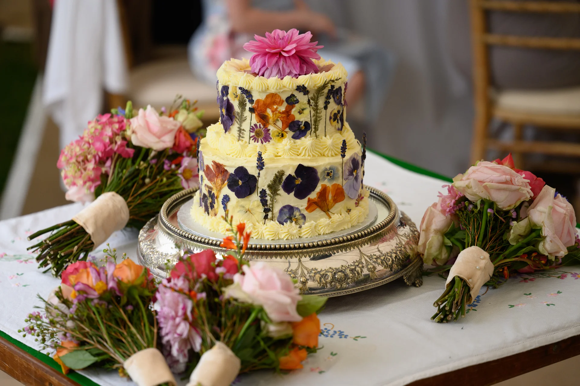 A stunning two-tier buttercream wedding cake decorated with pressed edible flowers sits on an ornate silver cake stand, surrounded by several colourful hand-tied floral bouquets on a table.