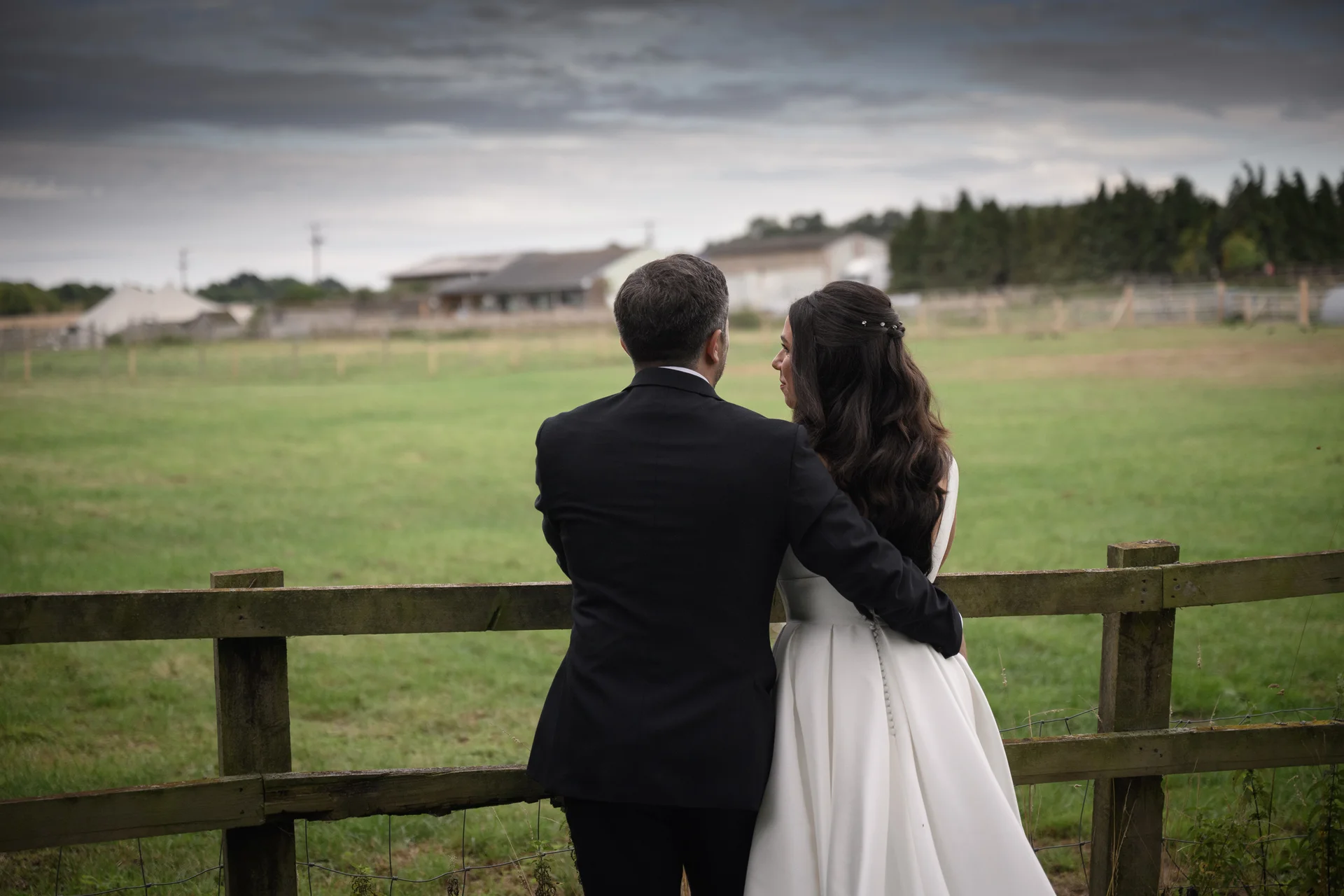 A bride and groom stand with their backs to the camera, leaning against a wooden fence and looking out over a green farm landscape under a dramatic cloudy sky.