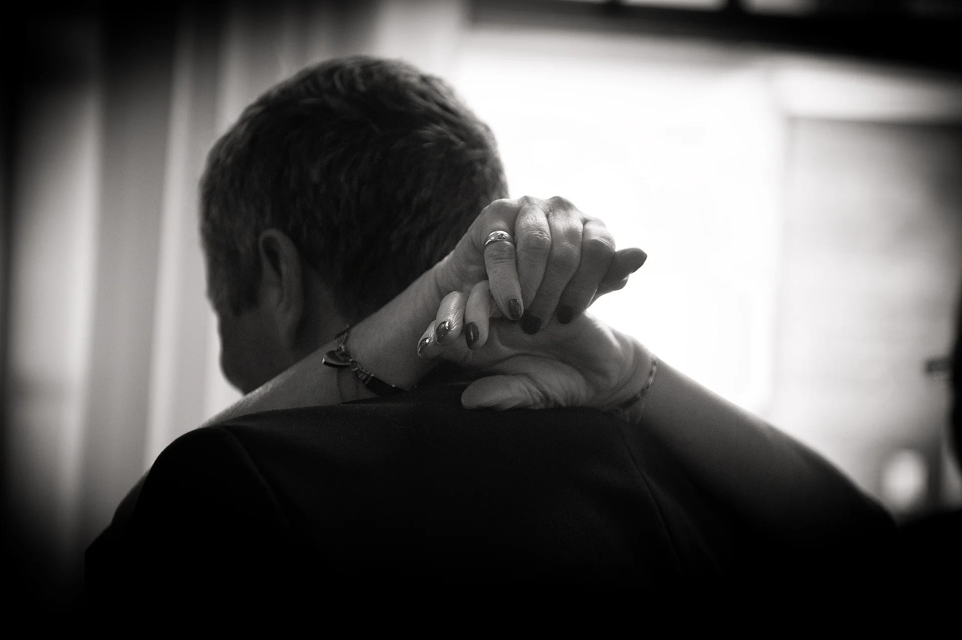 An intimate black and white back-view photograph capturing a couple embracing closely while dancing, with hands gently resting on shoulders and wedding bands visible.