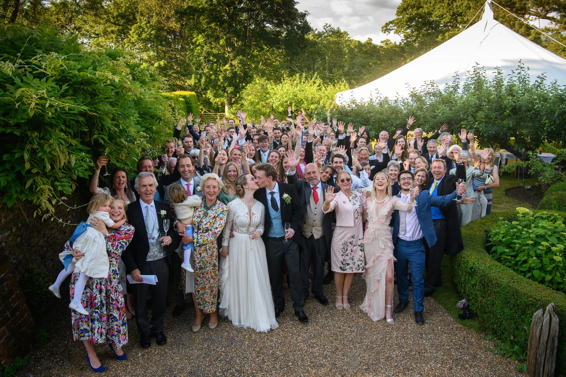 A large wedding crowd raises their glasses and hands in celebration on a gravel path outside a white marquee during a sunny garden reception.