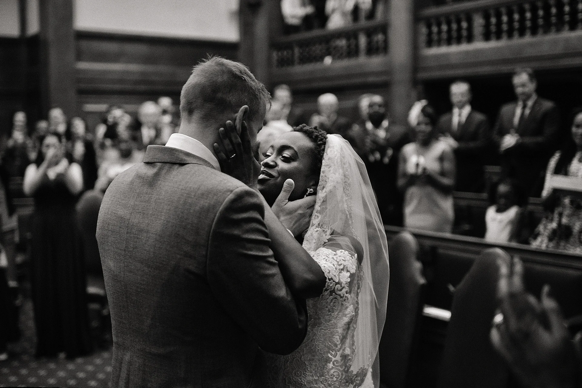 An interracial newlywed couple shares a tender moment embracing inside a historic church while wedding guests applaud in the background.